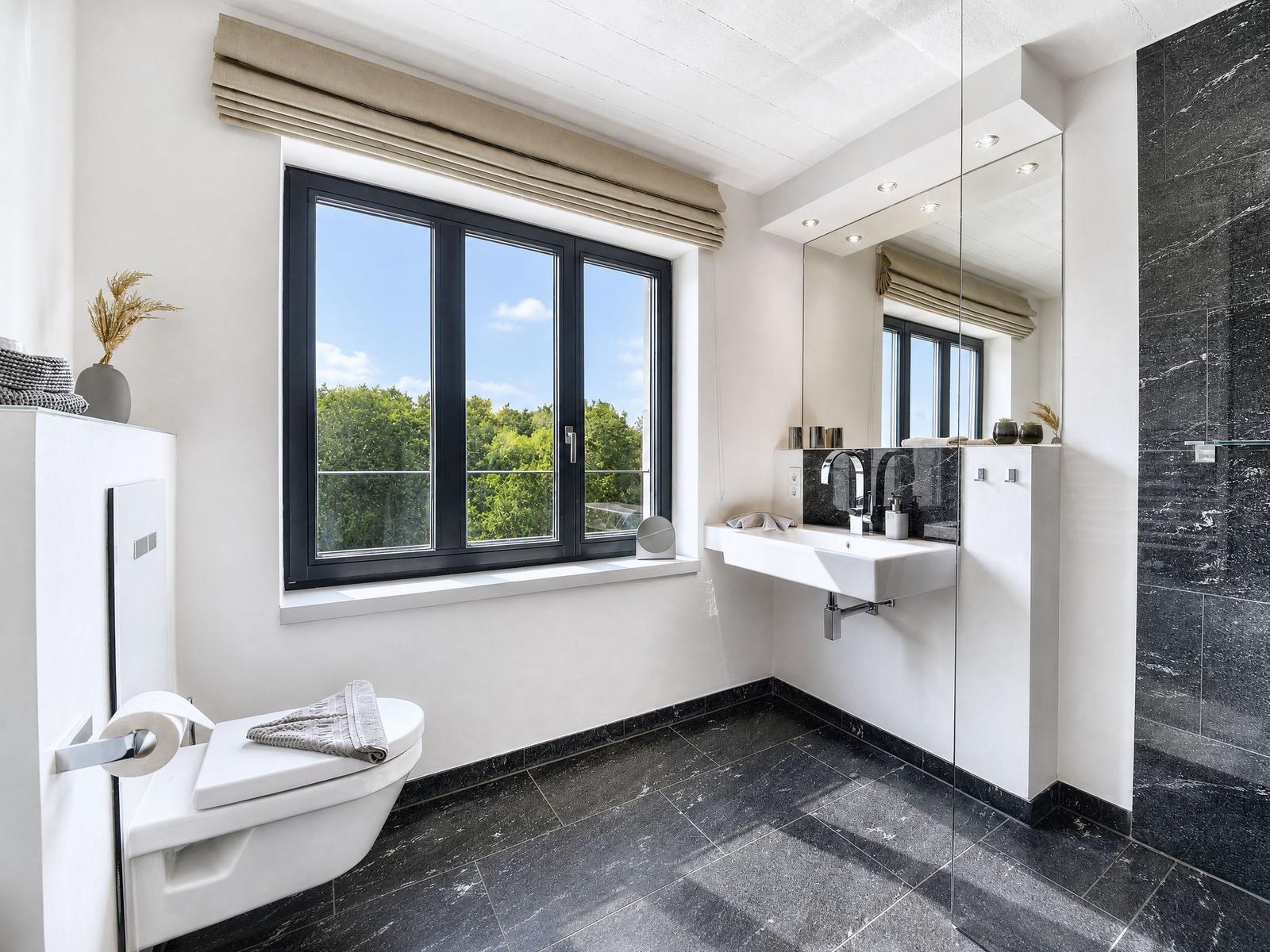 Badezimmer mit Duschbereich, Waschbecken und Toilette. Große Fenster mit Blick auf Grün.