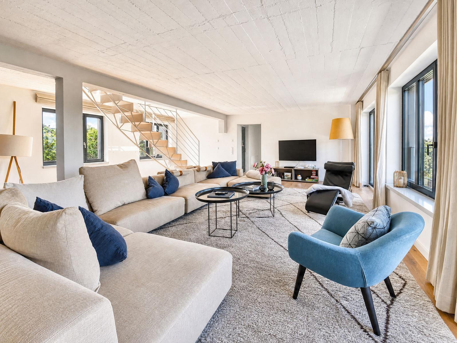 Wohnzimmer mit Sofa, Sessel und Fernseher. Treppe führt zur oberen Etage.