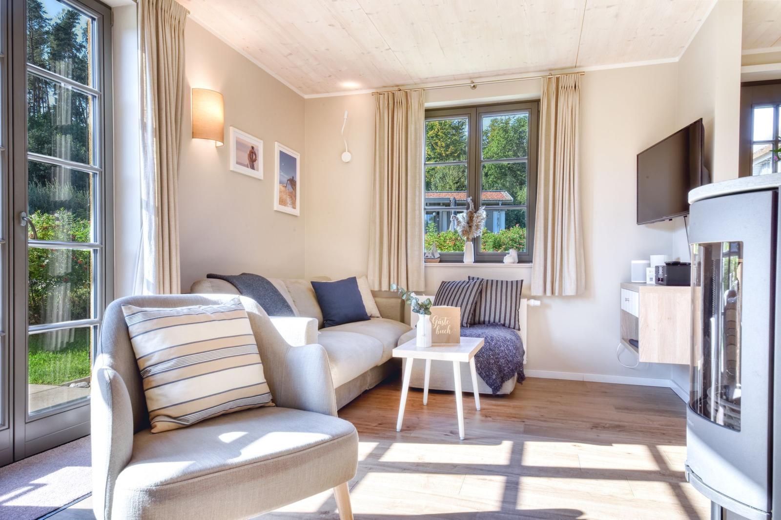 Wohnzimmer mit Sofa, Sessel und Fernseher. Holzboden und große Fenster mit Blick auf Garten.