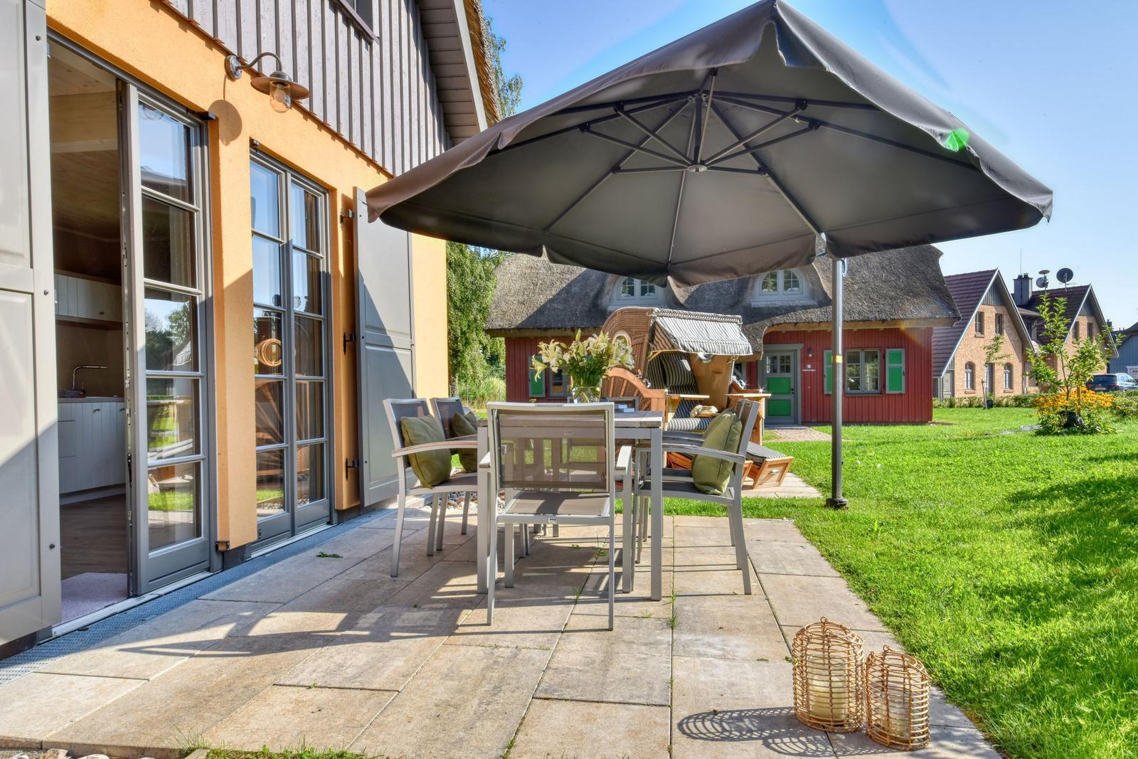 Terrasse mit Tisch, Stühlen und Sonnenschirm. Hintergrund: grüner Rasen und Häuser.