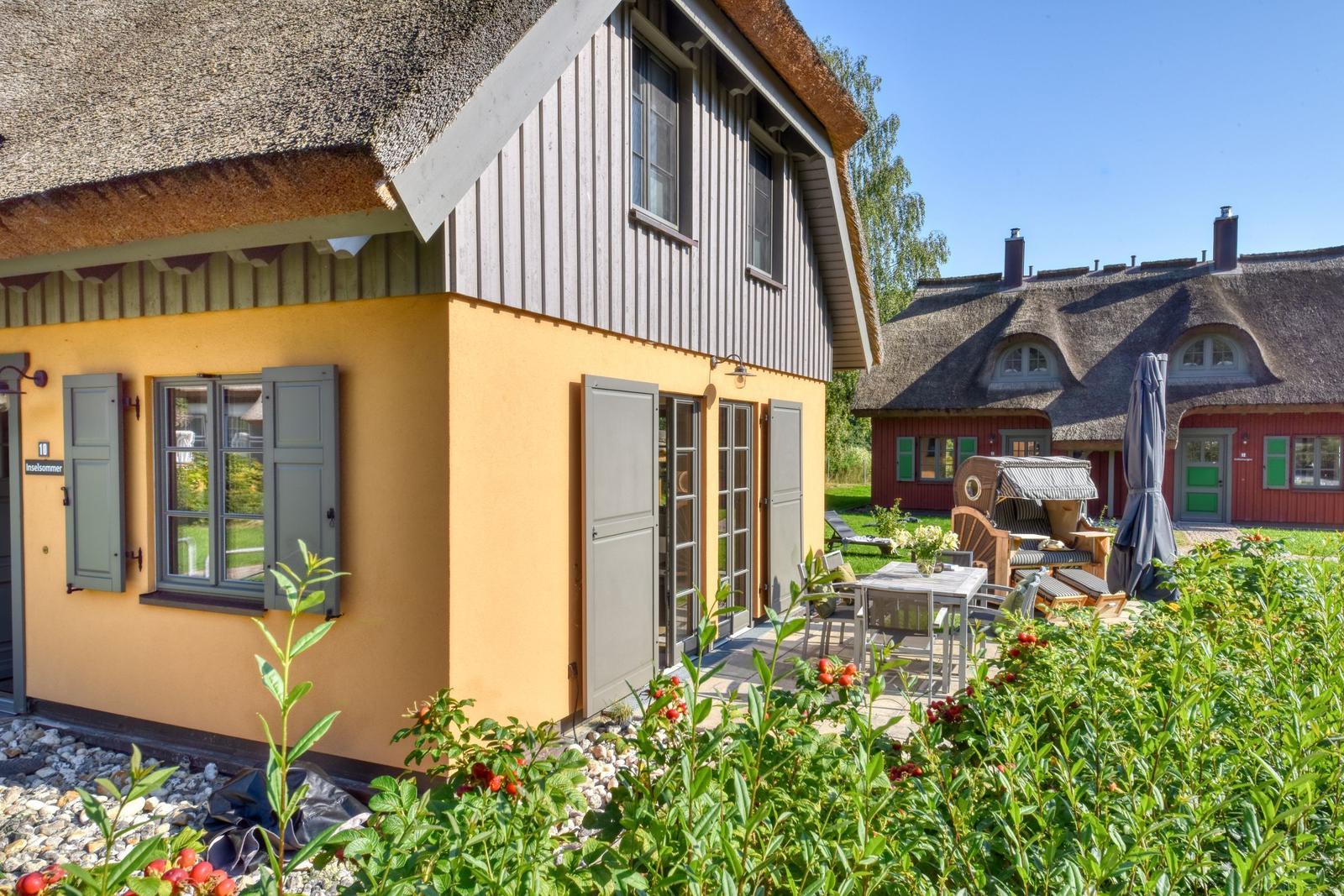 Haus mit Strohdach, Terrasse und Garten. Im Hintergrund ein weiteres Haus.