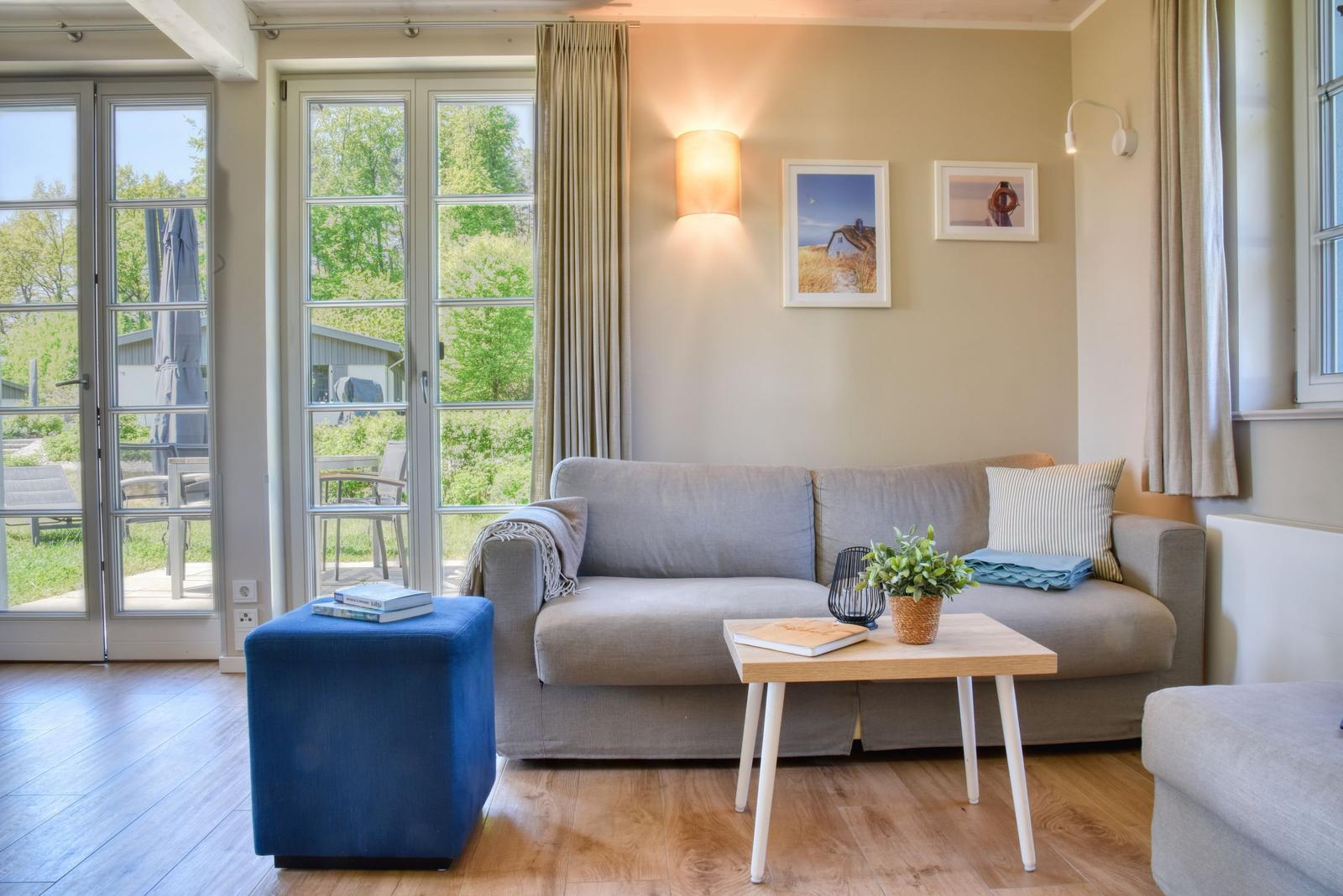Ein Wohnzimmer mit grauem Sofa, blauem Hocker und Holztisch. Durch Fenster ist ein Garten sichtbar.