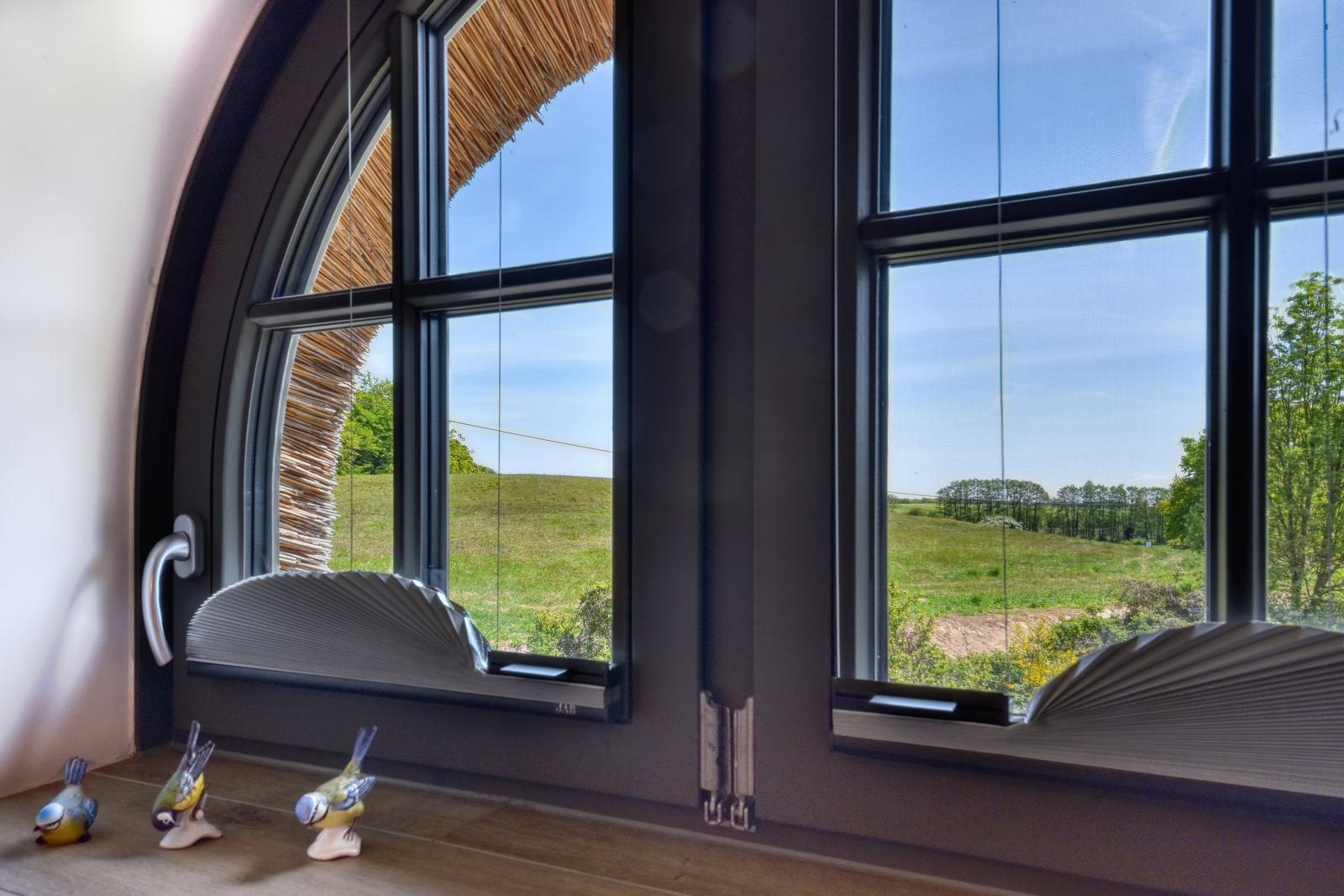 Großes Fenster mit Blick auf grüne Wiesen und einen Strohdachbereich.