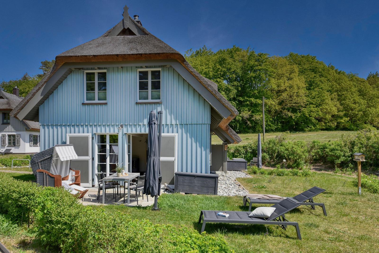 Blau gestrichenes Haus mit Dach aus Stroh, Terrasse mit Tisch und Stühlen, Liegestühle auf Rasen.