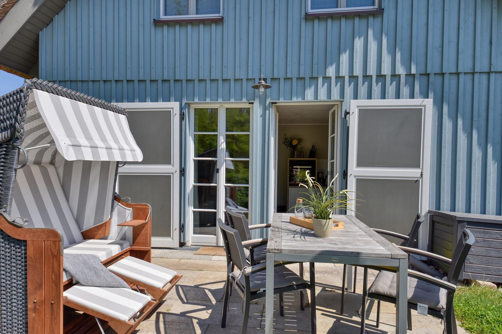 Terrasse mit Liege, Tisch und Stühlen vor blauem Haus mit offener Tür.