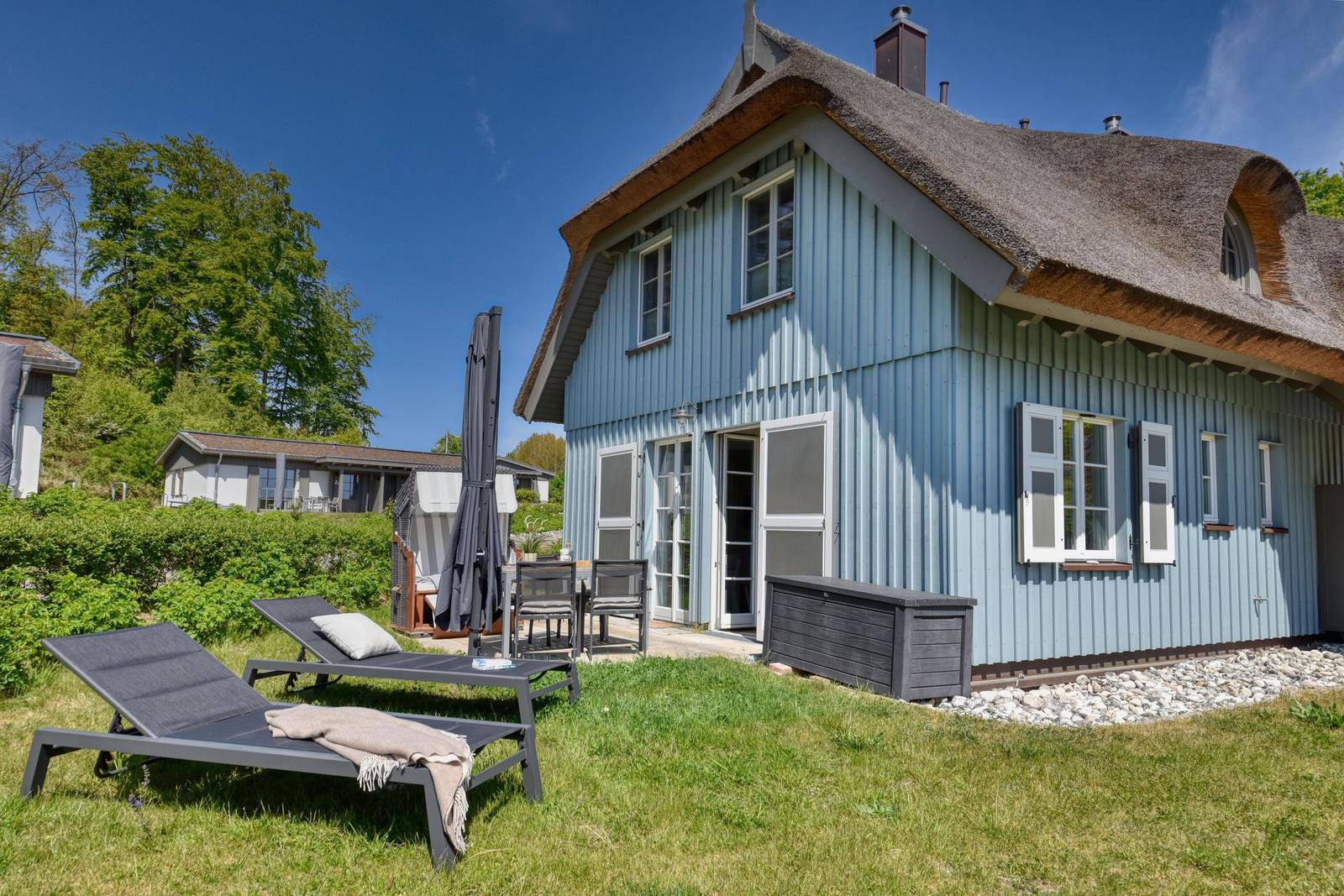 Blau gestrichenes Haus mit Dach aus Stroh, Gartenbereich mit Liegestühlen und Sonnenschirm.