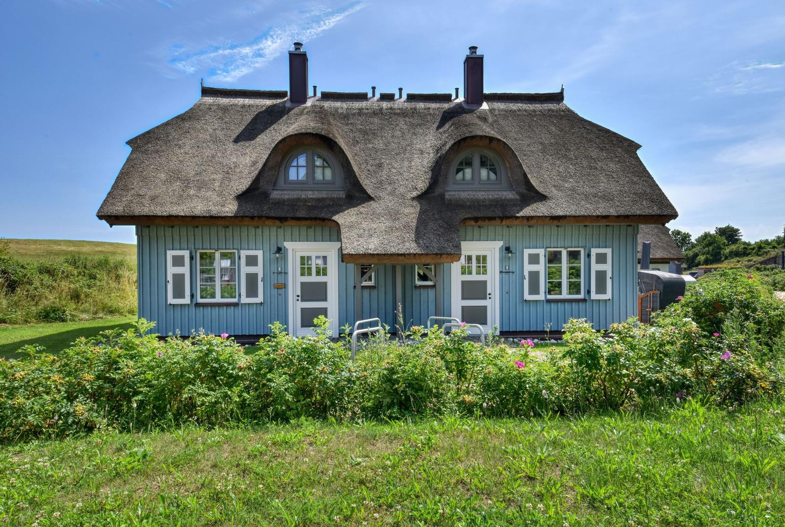 Blau gestrichenes Haus mit Strohdach und weißen Fenstern. Grünflächen und Blumen im Vorgarten.