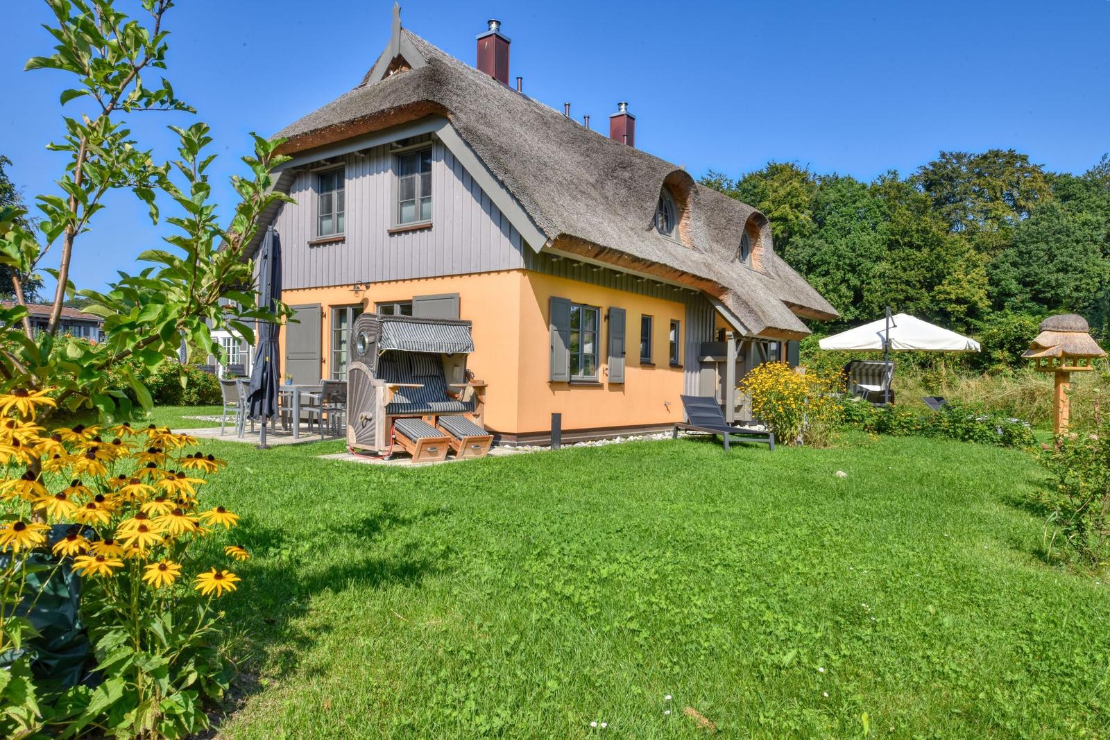 Haus mit Strohdach, Garten mit Liegen und Sonnenschirm. Blumen und Gras im Vordergrund.