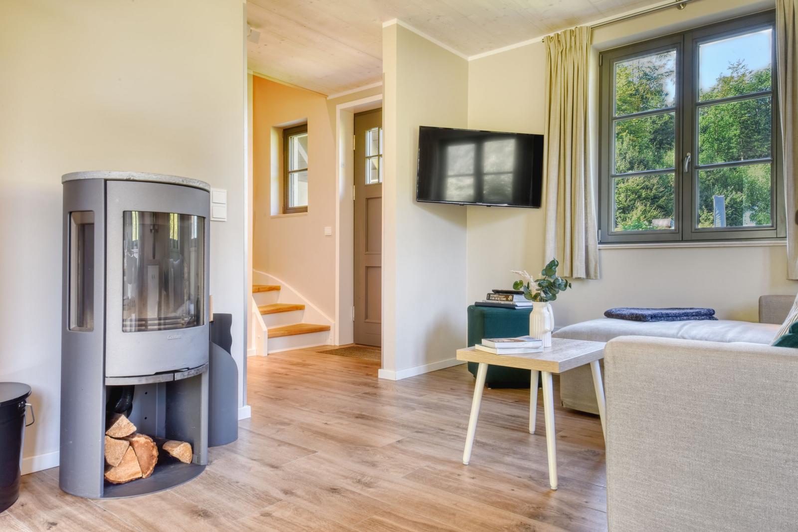 Wohnzimmer mit Kamin, TV und Blick auf Wald. Holzdielenboden und Fenster mit Vorhängen.