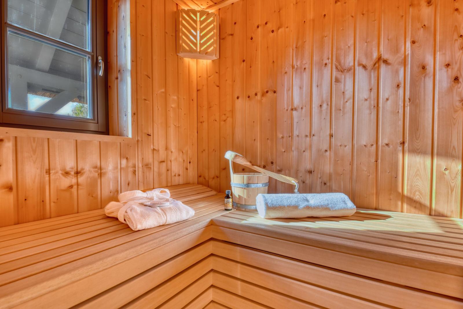Holzsauna mit Sitzbänken, Fässchen und Handtüchern. Fenster und Wandbeleuchtung sichtbar.