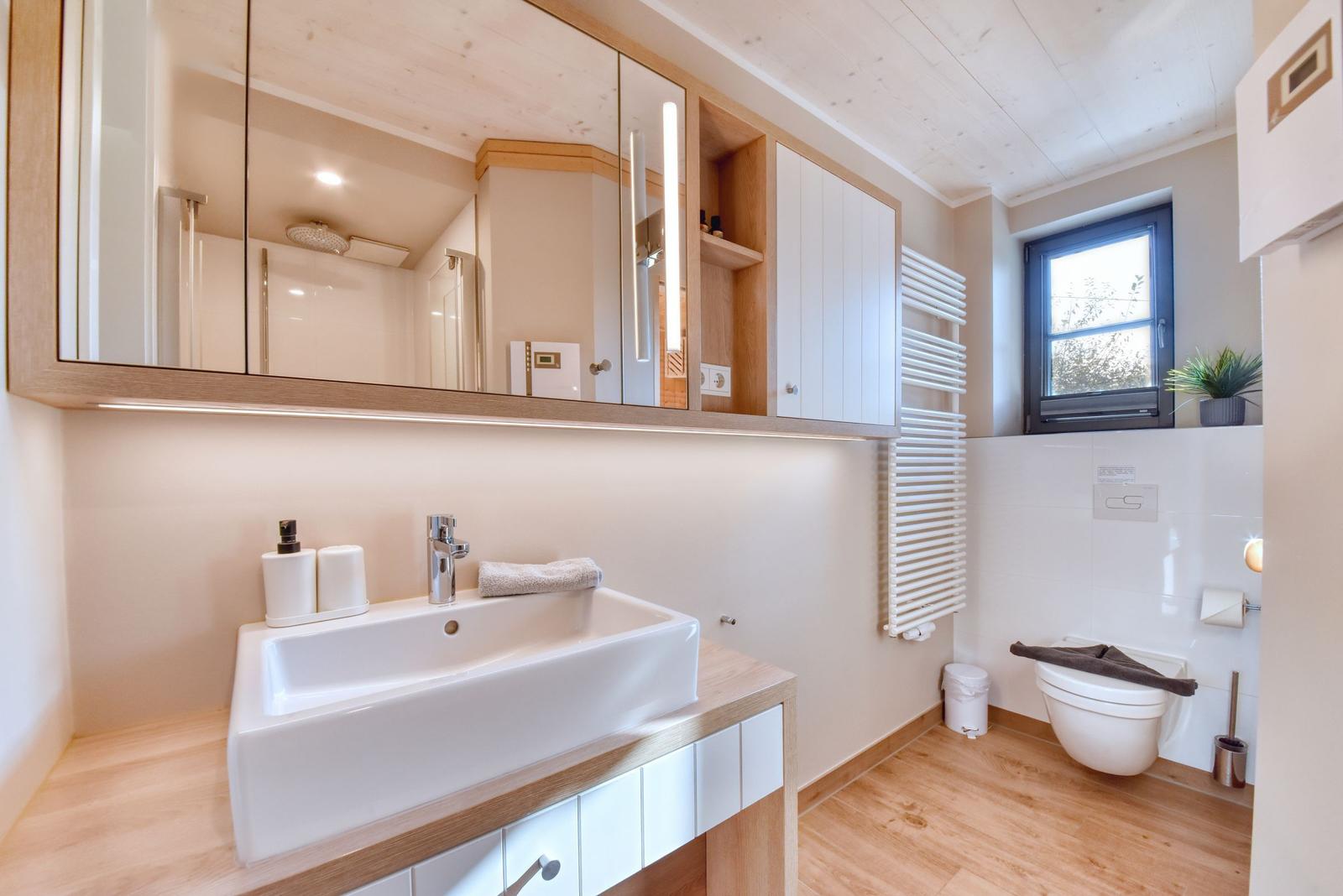 Badezimmer mit Waschbecken, Spiegelschrank und Toilette. Holzboden und Fenster sichtbar.
