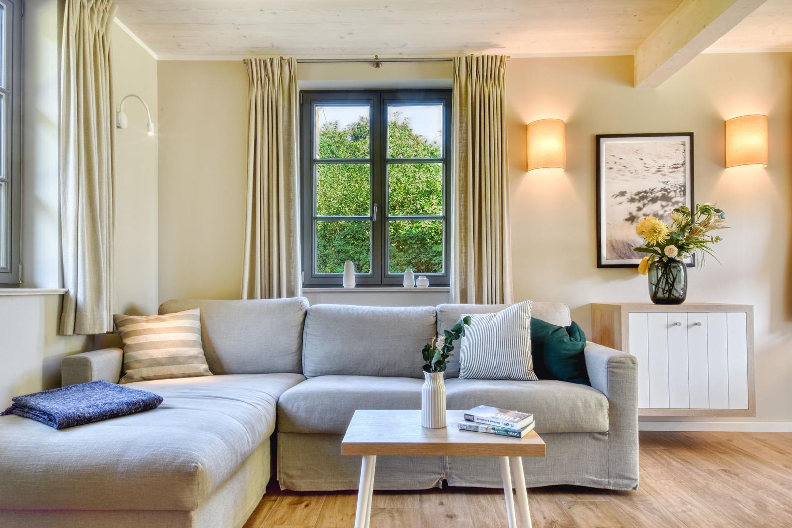 Ein Wohnzimmer mit grauem Sofa, Holztisch und Fenster mit Blick auf Bäume.
