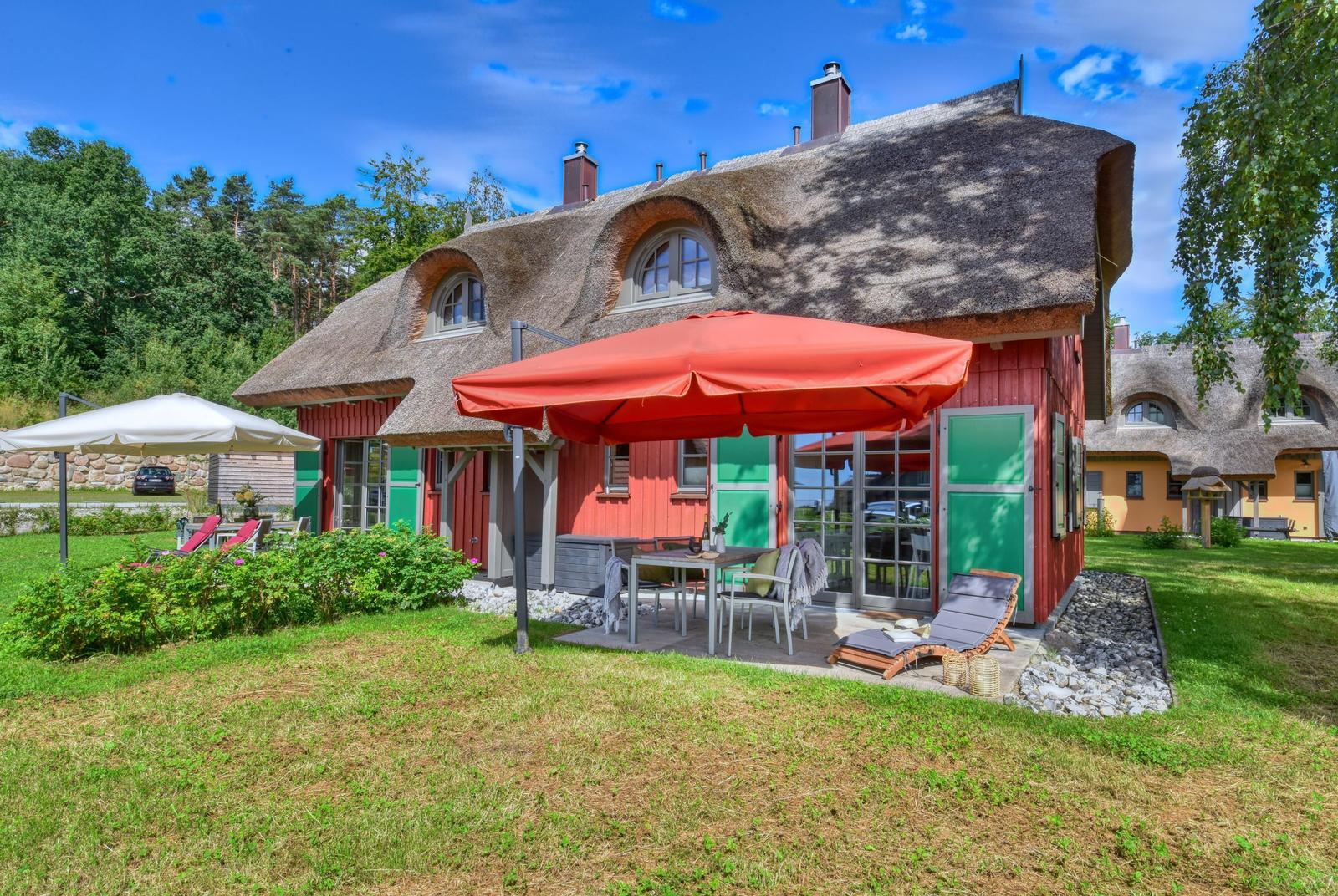 Haus mit Strohdach, Terrasse mit Tisch, Stühlen und Sonnenschirmen. Grün und Rot gestrichene Fassade.