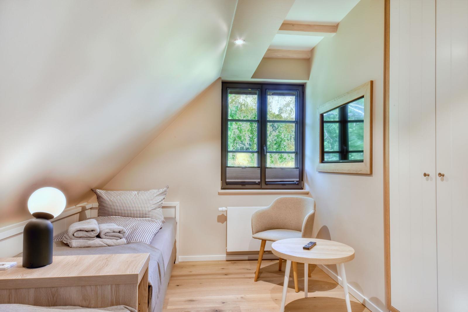 Schlafzimmer mit Dachschräge, Bett, Stuhl und Tisch. Fenster mit Blick auf Grün.