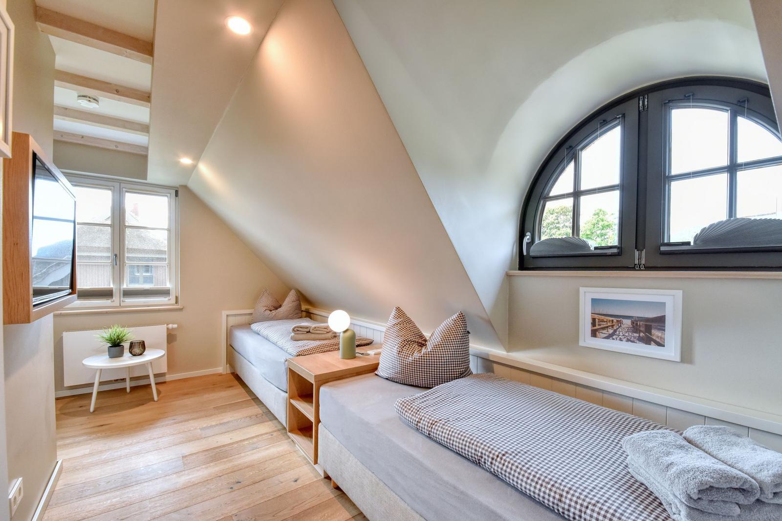 Zimmer mit zwei Betten, Holzboden und schrägen Wänden. Fenster und TV sichtbar.