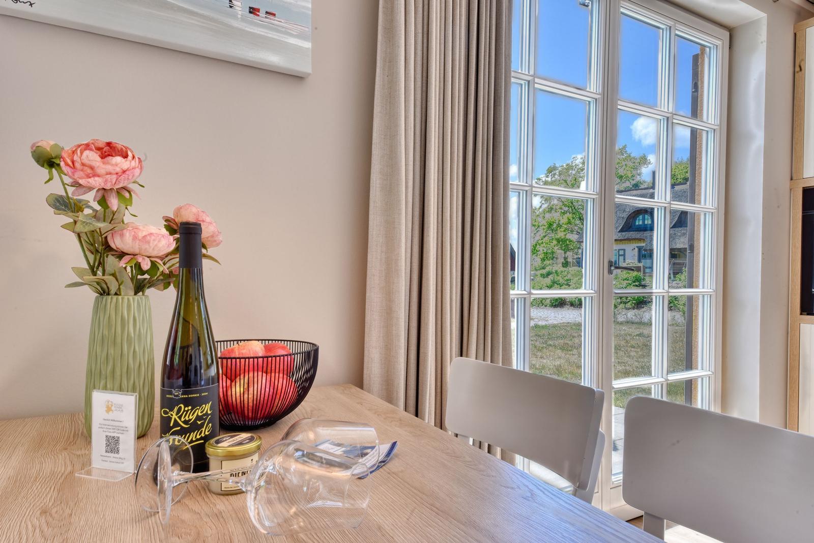 Esstisch mit Weinflasche, Blumen und Obst vor Fenster mit Blick auf Haus und Grün.