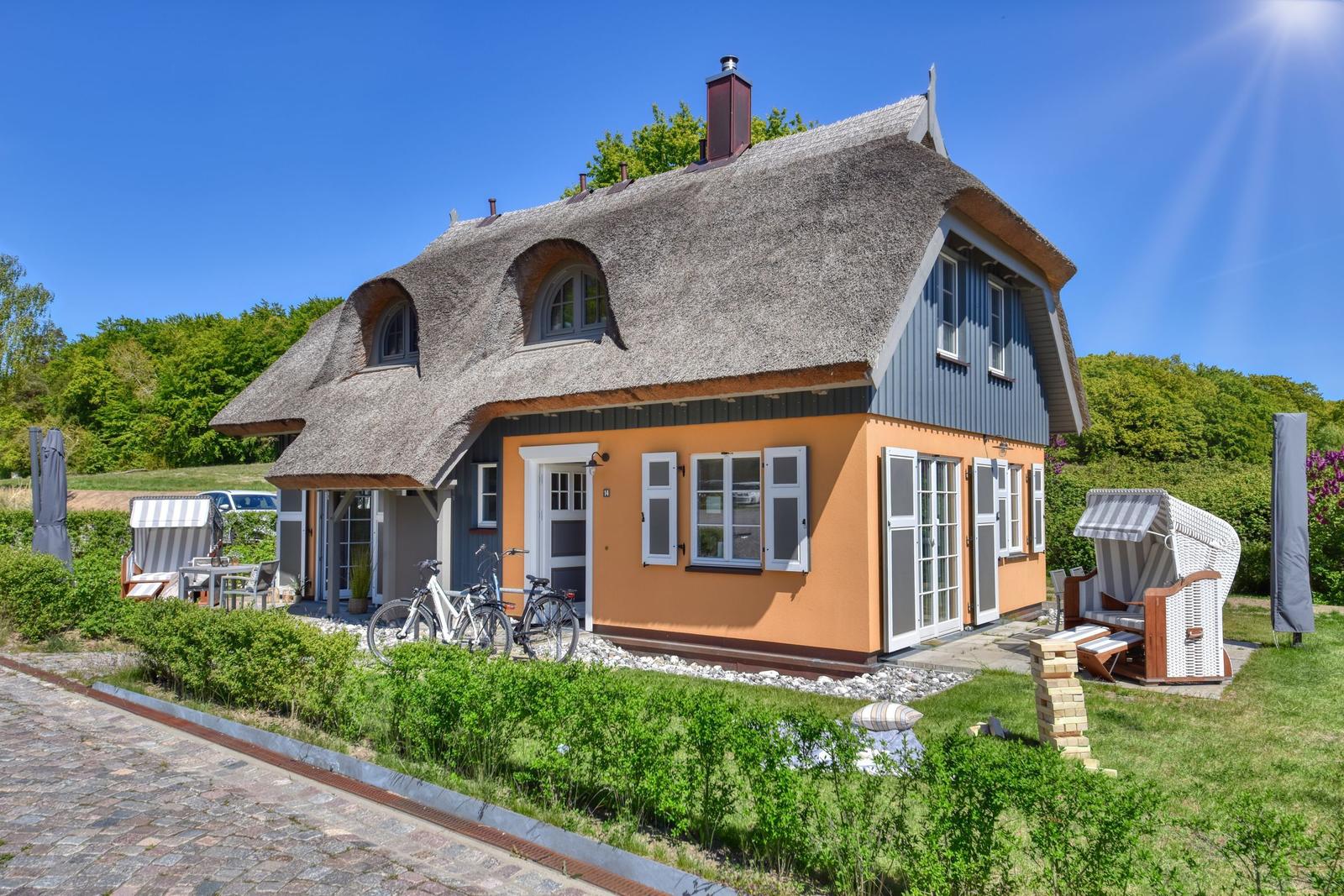 Haus mit Strohdach, orangefarbener Fassade und Gartenbereich mit Liegestühlen.