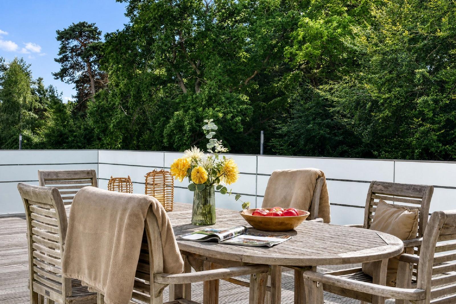 Holztisch mit Stühlen, Blumen und Äpfeln auf Terrasse mit Blick auf Bäume.