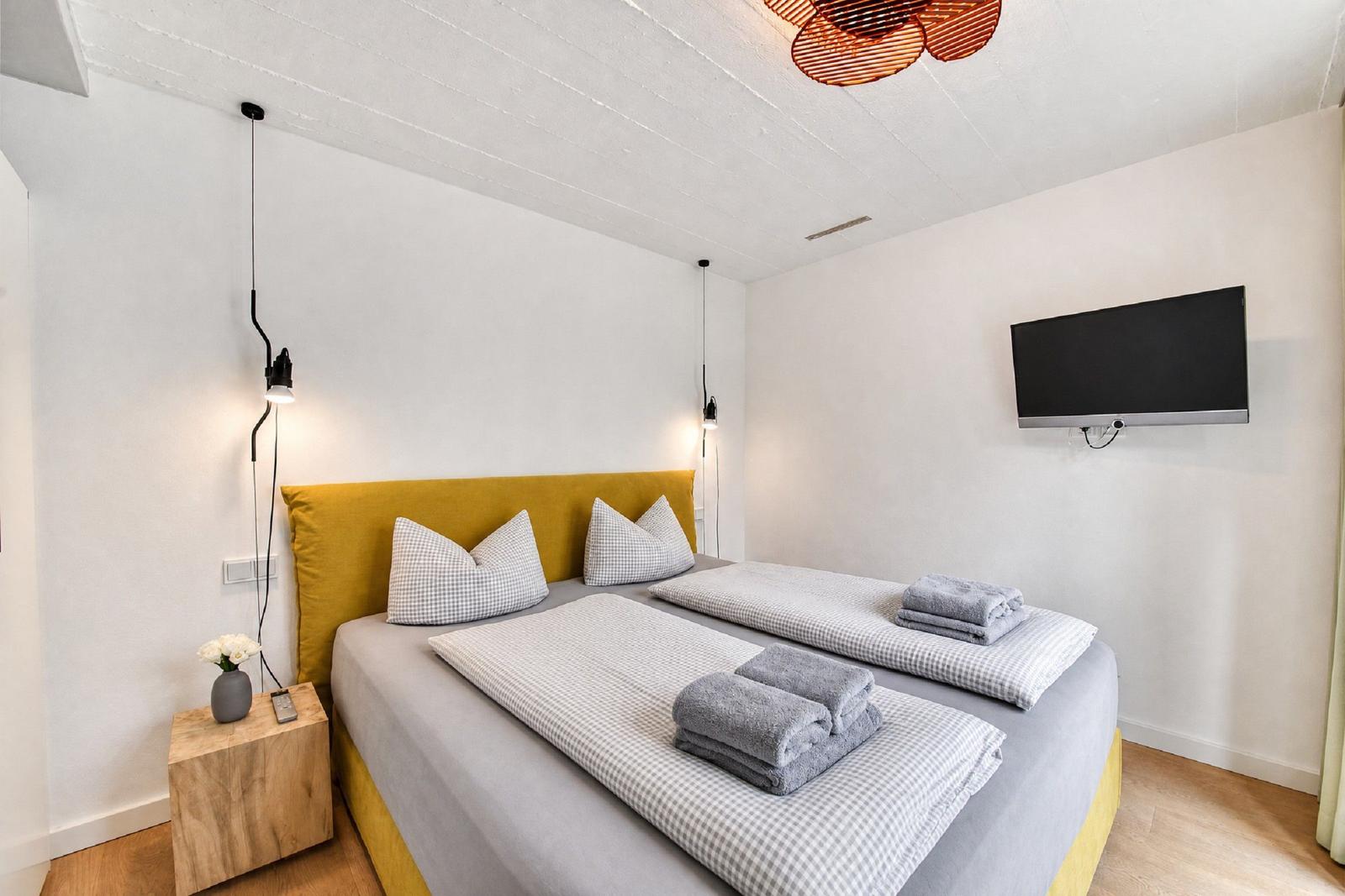 Schlafzimmer mit Doppelbett, gelbem Kopfteil, Fernseher und Nachttisch.