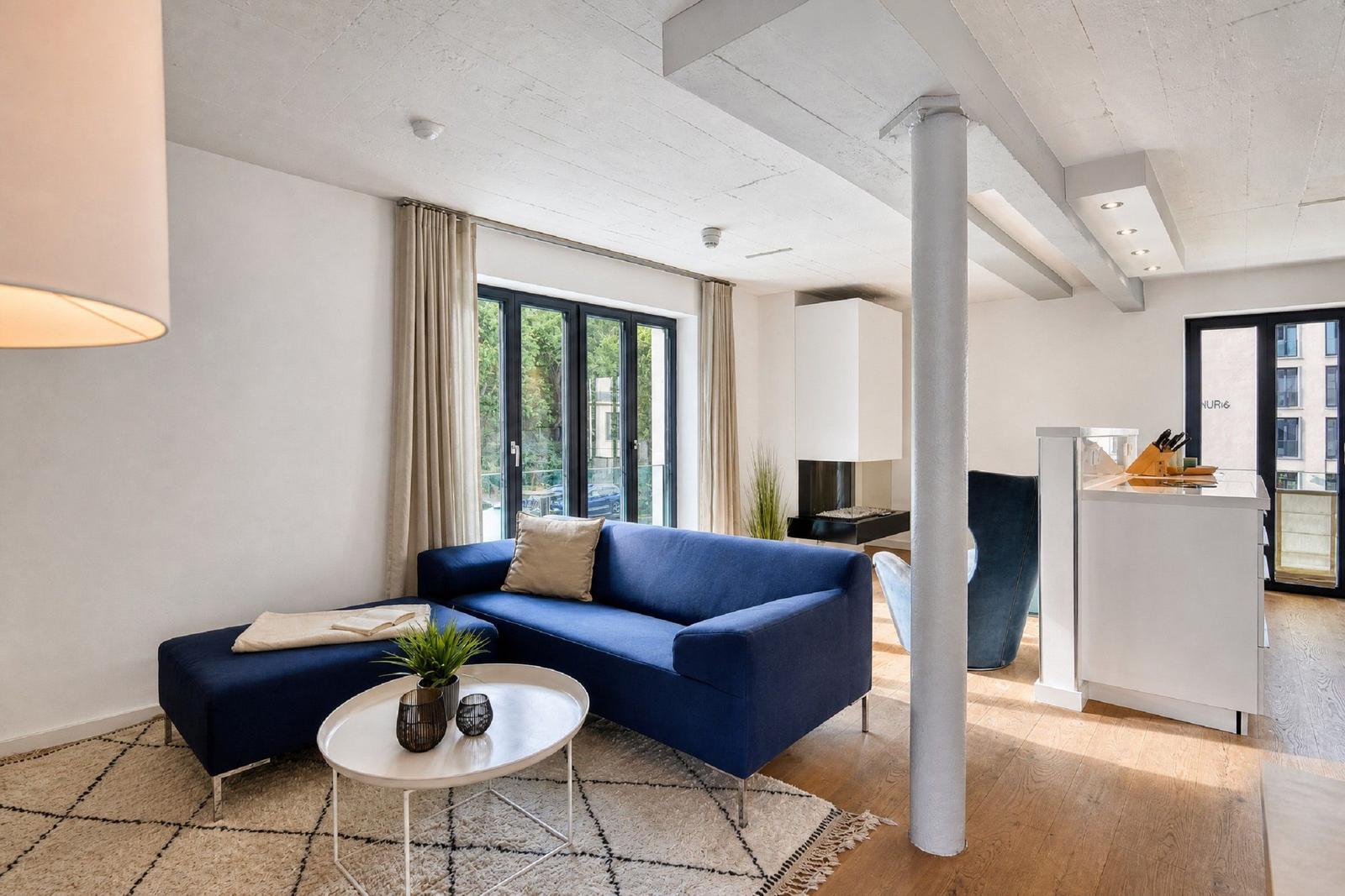 Ein Wohnzimmer mit blauem Sofa, weißem Tisch und Holzboden. Durchgehende Fensterfront mit Blick nach draußen.