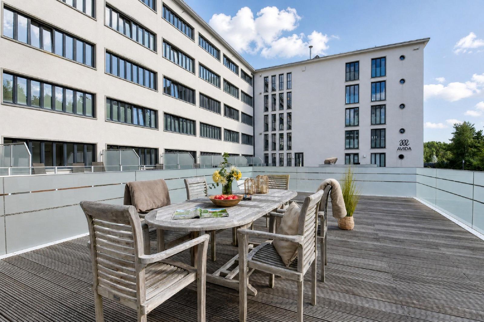 Terrasse mit Holztisch und Stühlen, Blick auf Gebäude mit Glasgeländer.