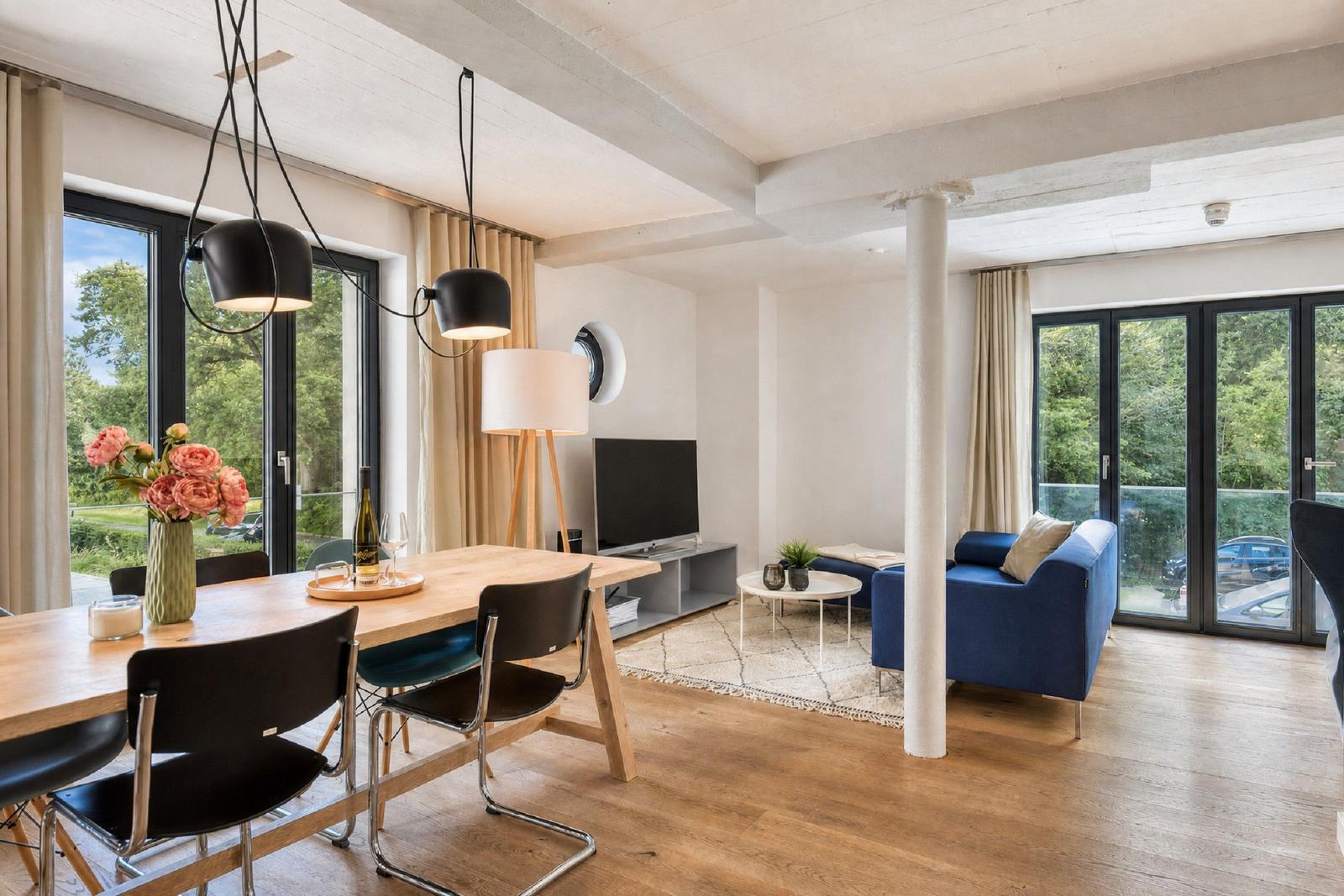 Wohn- und Essbereich mit Holzboden, blauem Sofa und großen Fenstern.