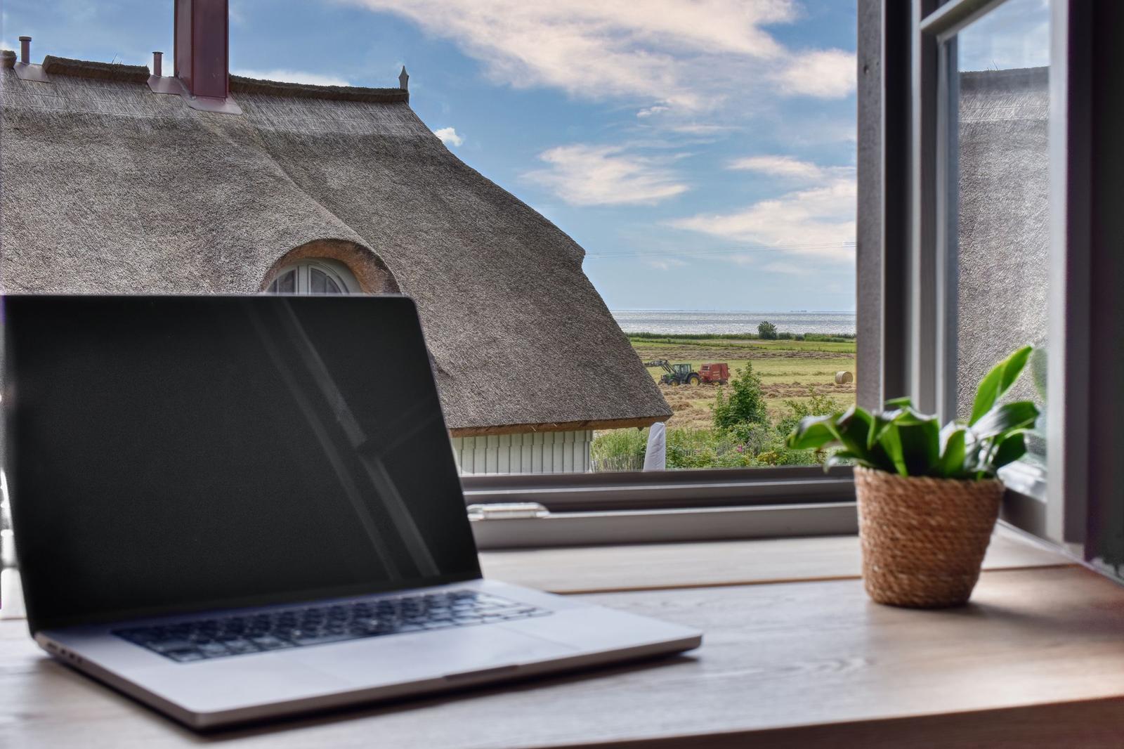 Laptop on table by window with view of thatched roof house and field.