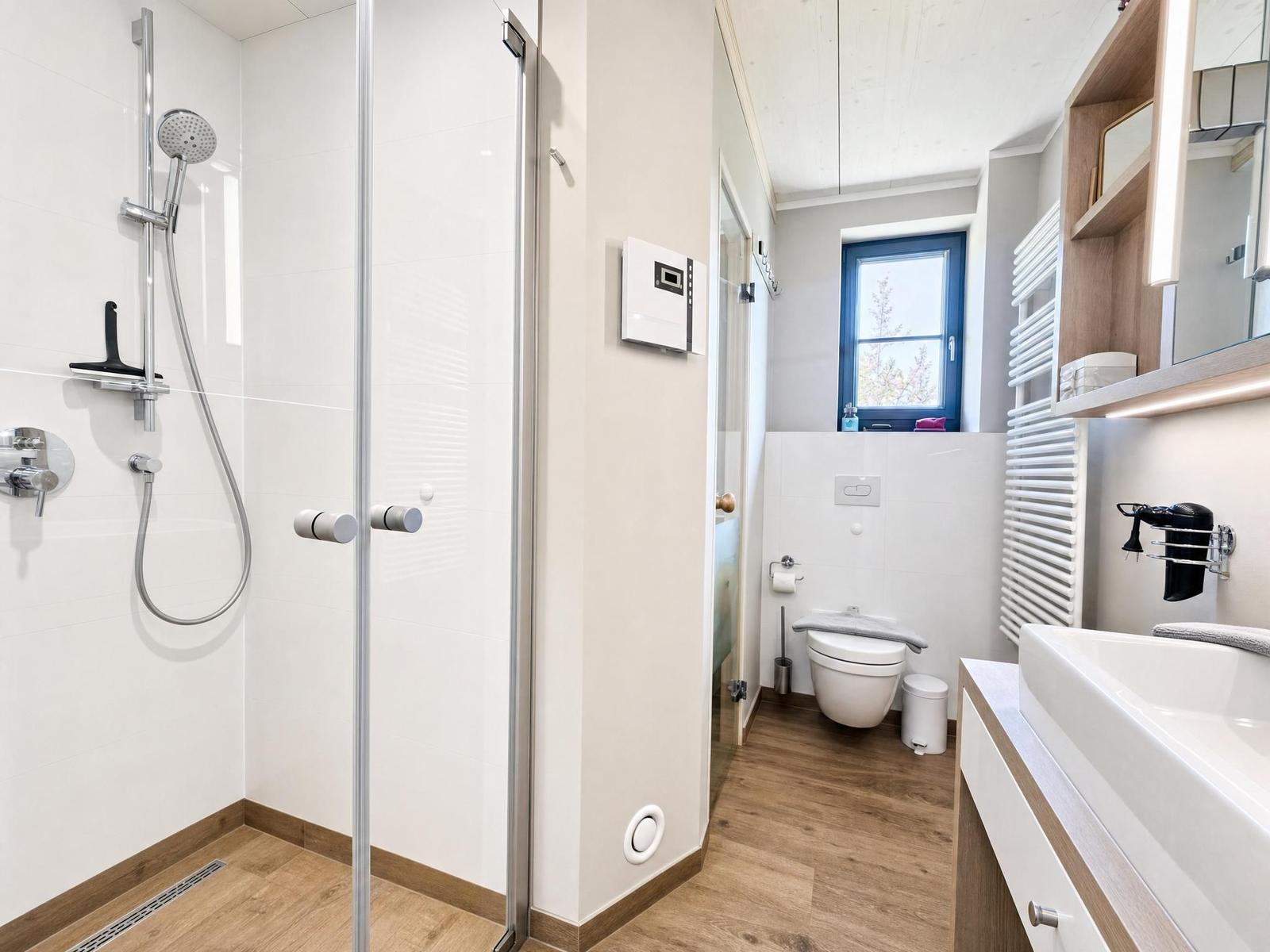 Bathroom with shower, toilet, and sink. Wooden floor and window with blue frame.
