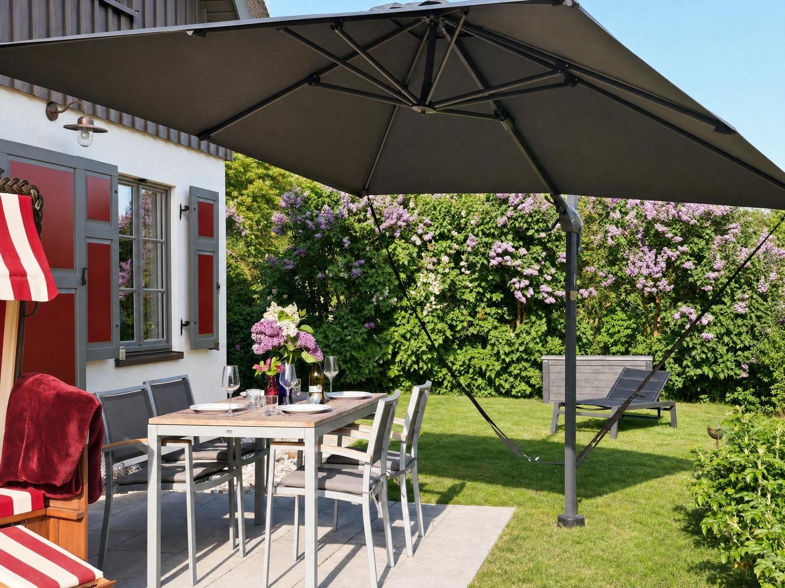 Terrace with table, chairs, and umbrella. Background: green garden with flowering bushes.