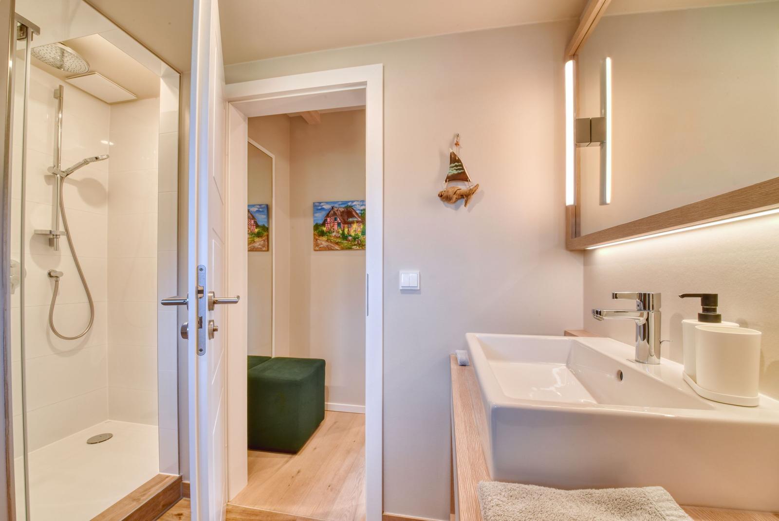 Badezimmer mit Dusche, Waschbecken und Spiegel. Durchgang zu einem Raum mit grünem Sitzkissen.