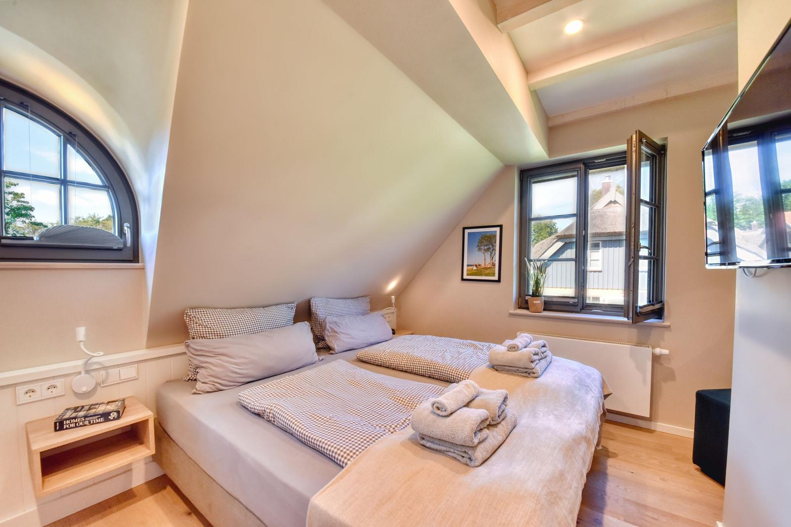Double bed with towels, windows, and TV in a bedroom with sloped ceilings.