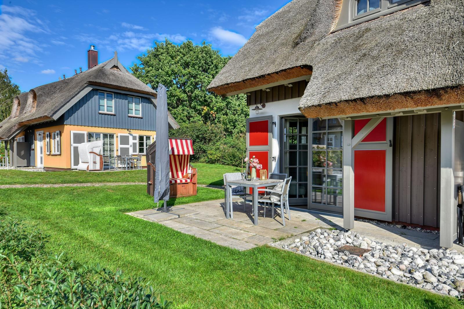 House with thatched roof, patio and garden. Red doors and wooden furniture.