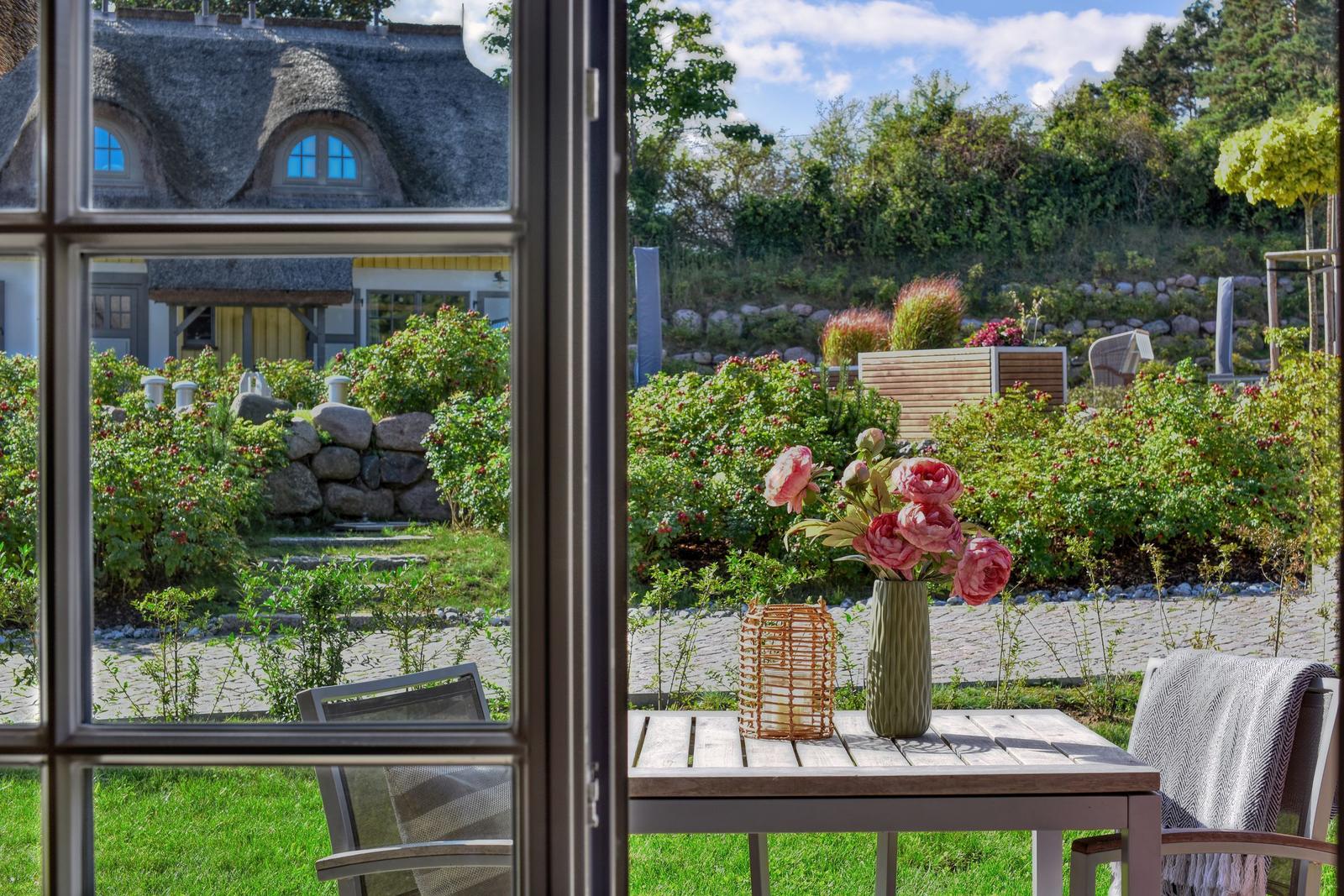 View through a window onto a garden with flowers and a villa with thatched roof.
