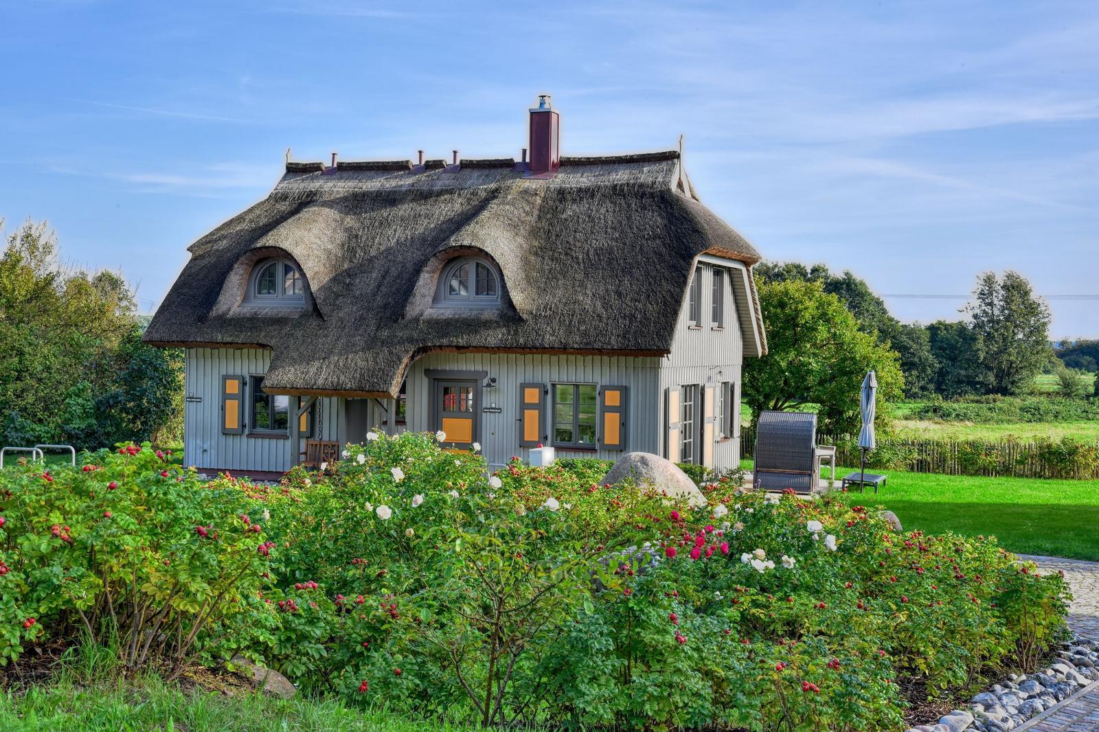 Haus mit Strohdach, Rosenbeet und Garten. Terrasse mit Liegestuhl und Sonnenschirm.