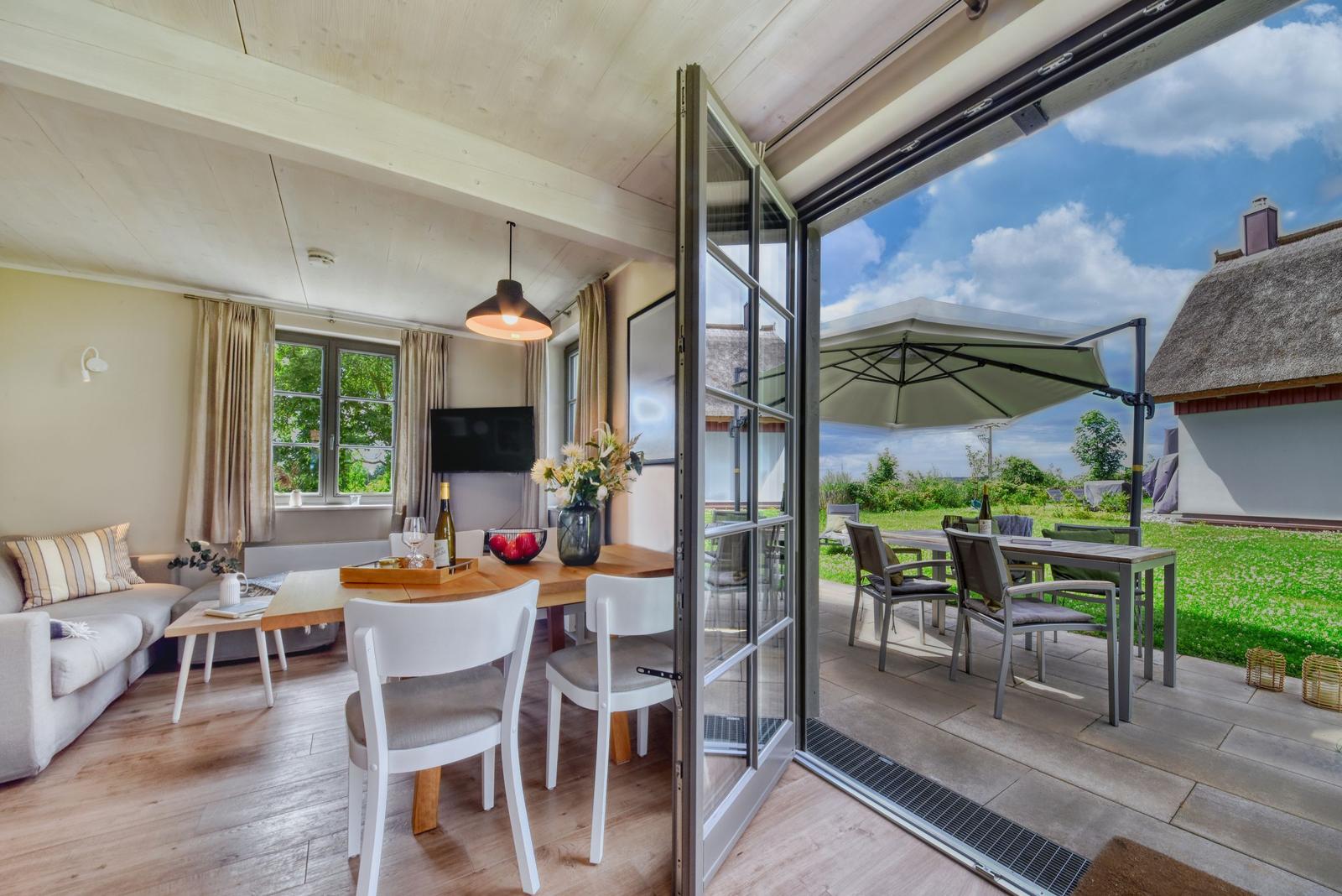 Wohn- und Essbereich mit Terrasse, Garten und Sitzgelegenheiten. Holzfußboden und große Fenster.
