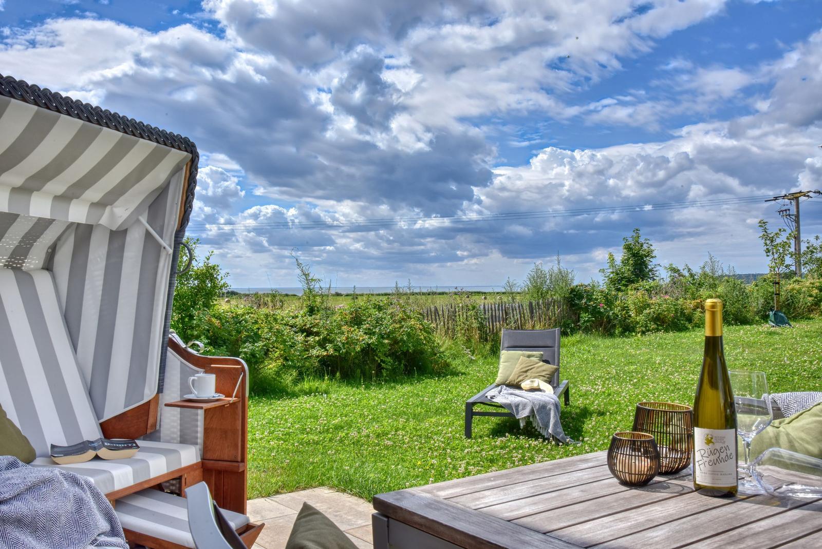 Terrasse mit Liegestuhl und Strandkorb. Tisch mit Wein und Kerzen. Blick auf Grünfläche und Horizont.