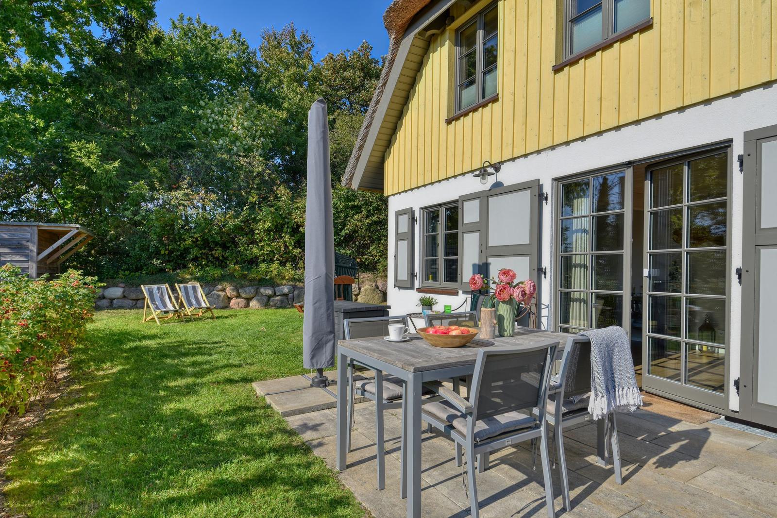 Haus mit Terrasse, Tisch, Stühlen und Garten. Außentisch mit Blumen und Obst.