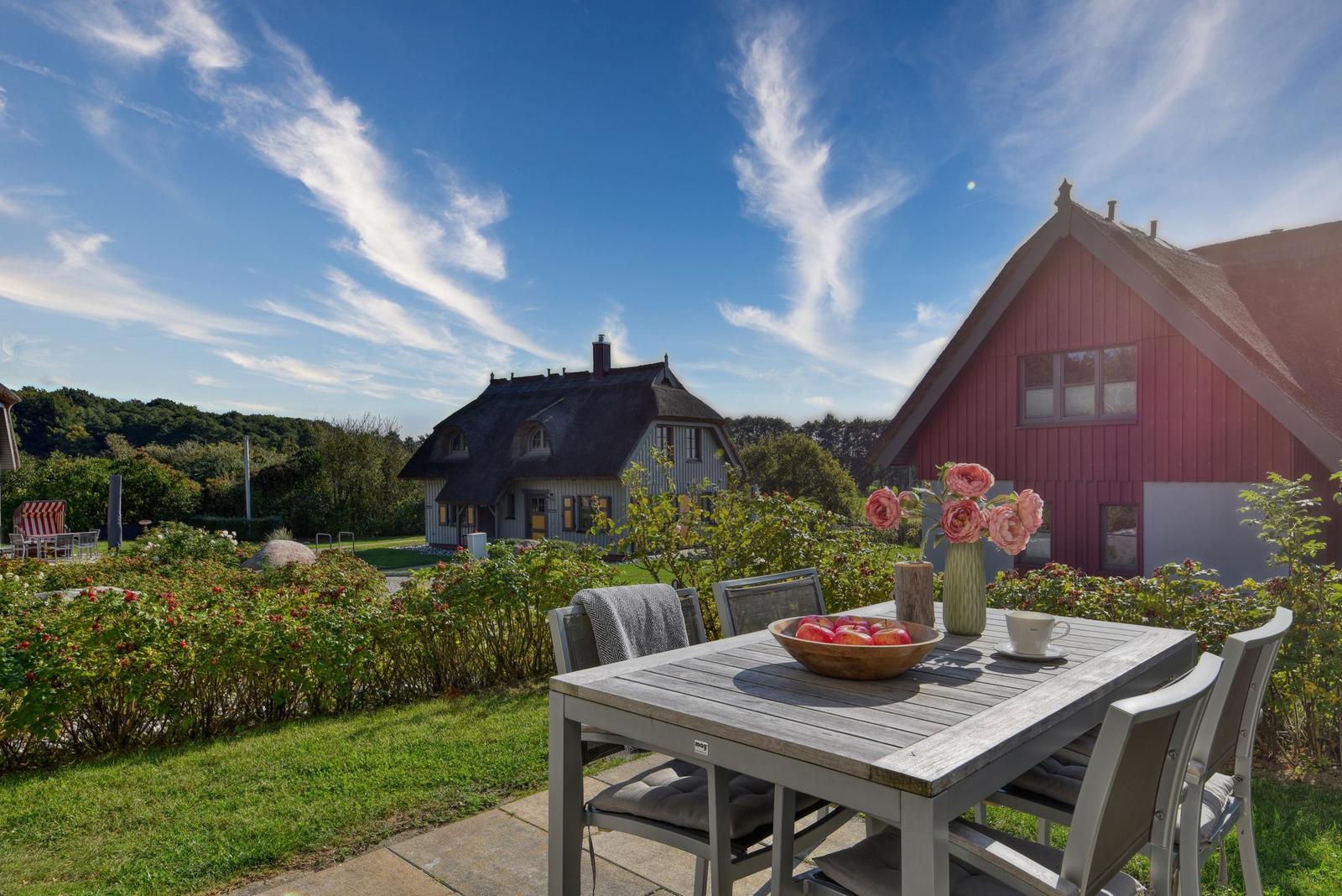 Terrasse mit Tisch und Stühlen. Im Hintergrund rotes und graues Haus mit Garten.