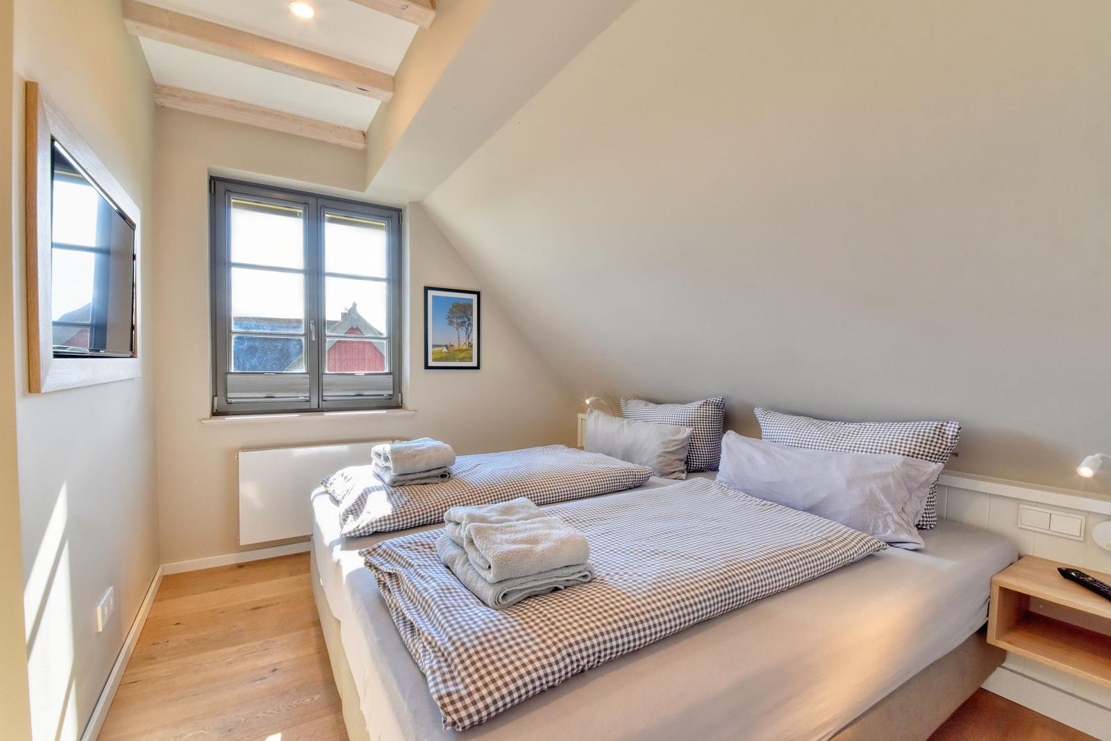 Schlafzimmer mit Doppelbett, Fenster und Holzboden. An der Wand hängt ein Fernseher.
