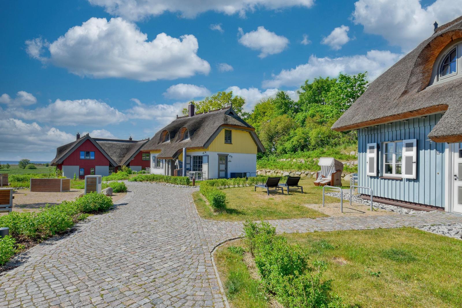 Three houses with thatched roofs stand along a paved road with garden and loungers.