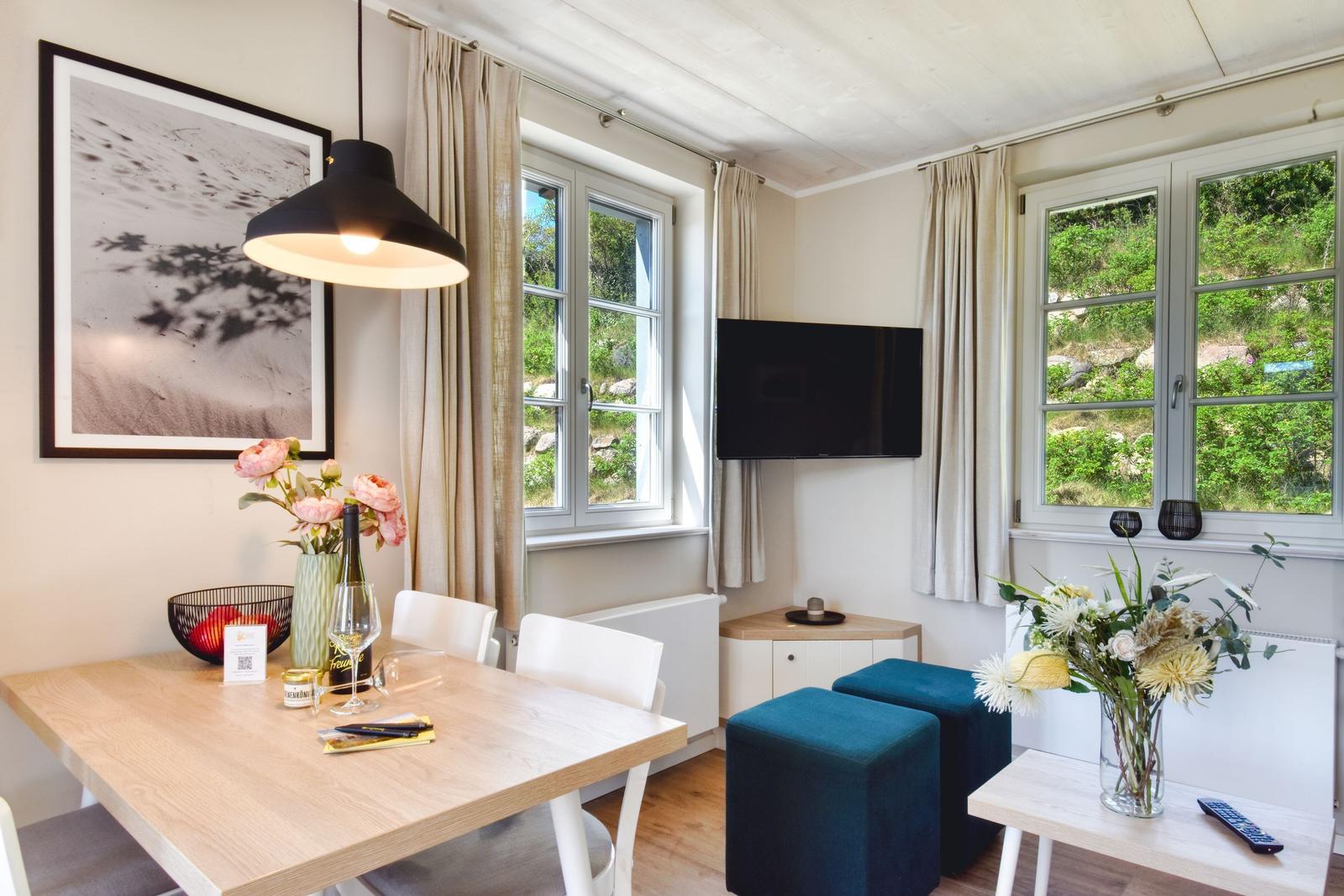 Dining area with table, chairs, and windows. TV and flowers are visible.