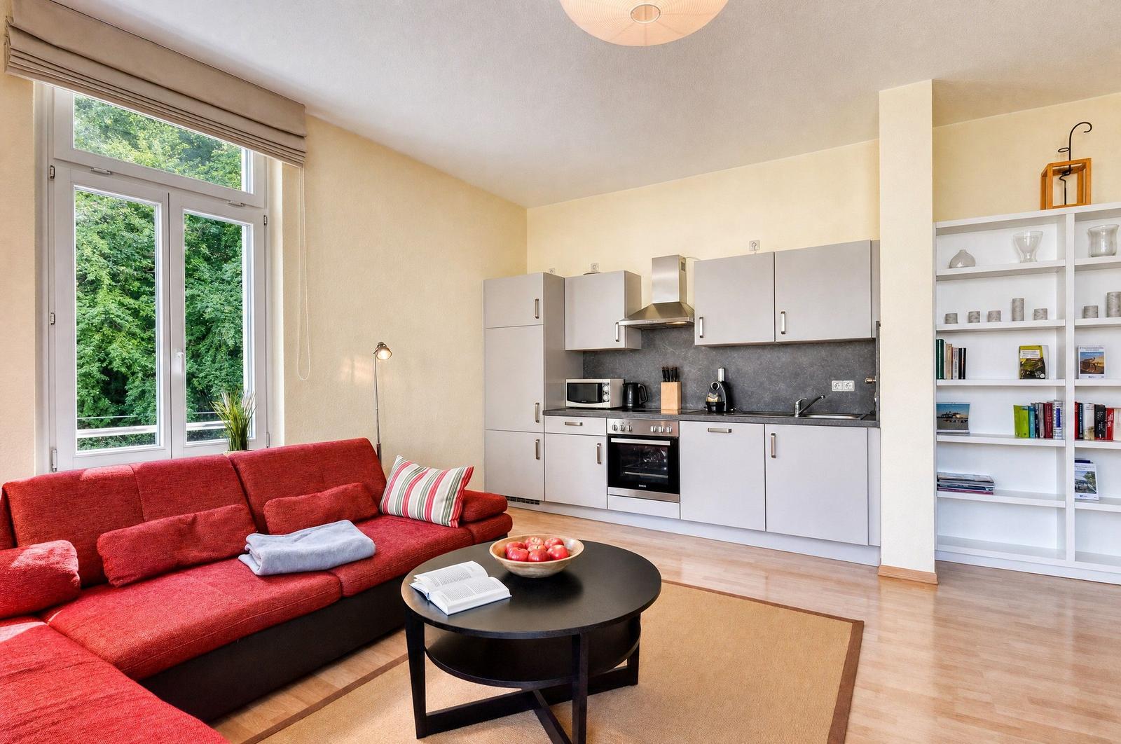 Wohnbereich mit rotem Sofa, Küchenzeile und Bücherregal. Fenster mit Blick auf Bäume.