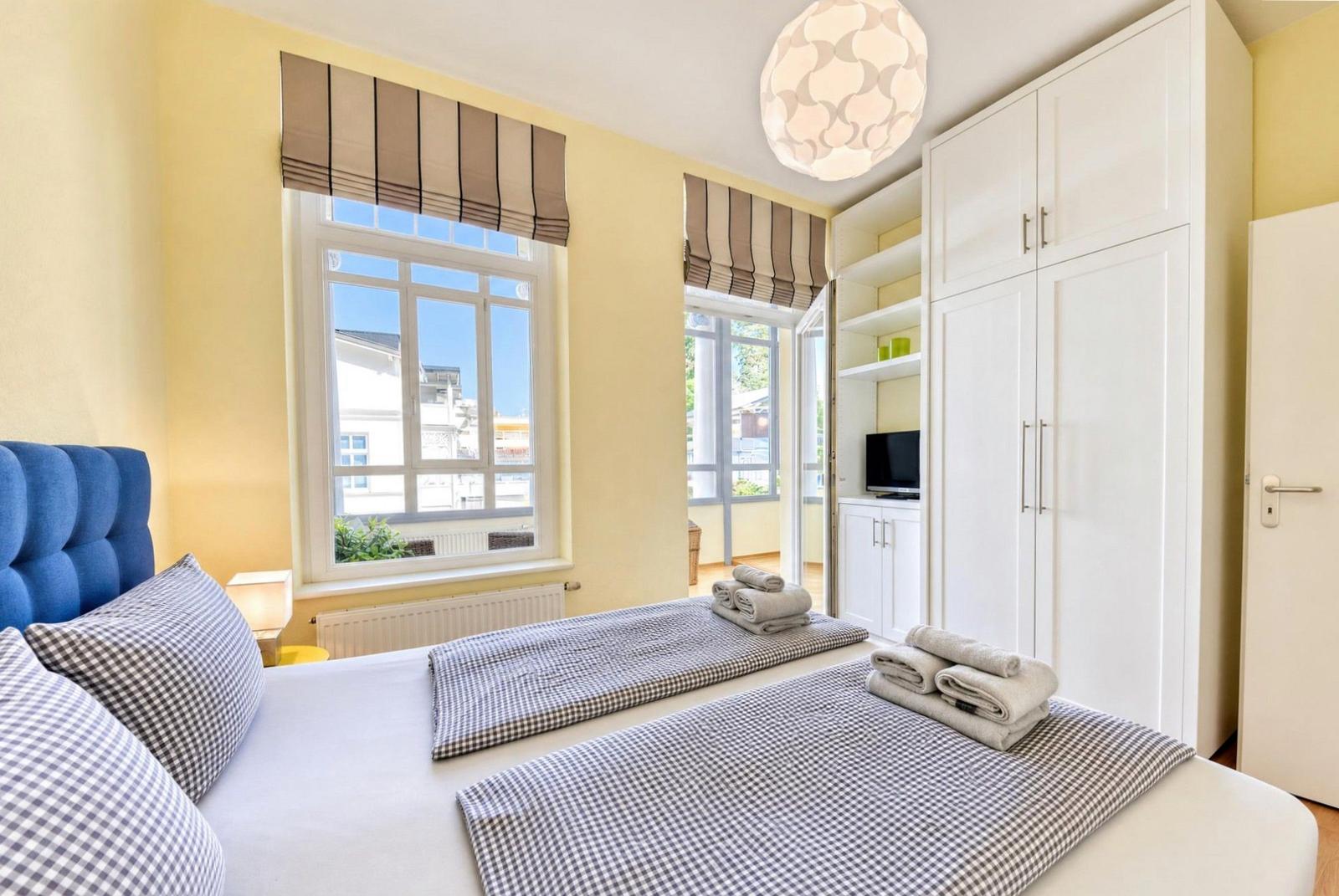Schlafzimmer mit blauem Kopfteil, weißen Wänden und zwei Fenstern. Weiße Schränke und ein Bett mit Handtüchern.