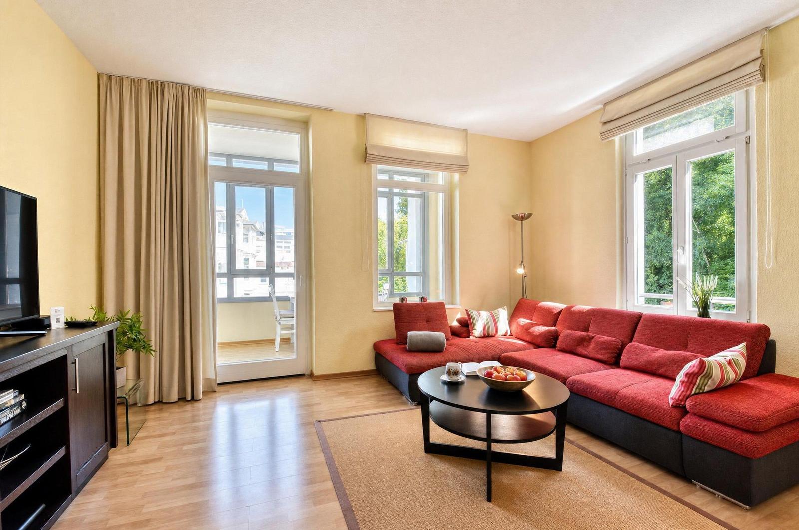 Ein Wohnzimmer mit rotem Sofa, Holzboden und großen Fenstern mit Blick auf grüne Bäume.