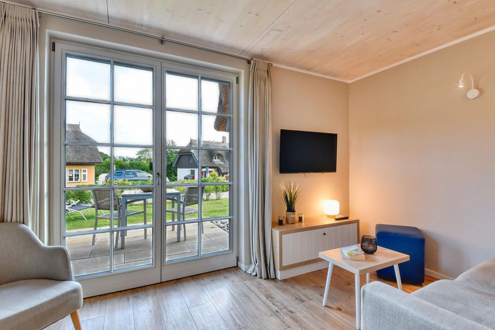 Wohnzimmer mit Fernseher, Tisch und Sitzgelegenheiten. Durchführung zur Terrasse mit Blick auf Häuser.