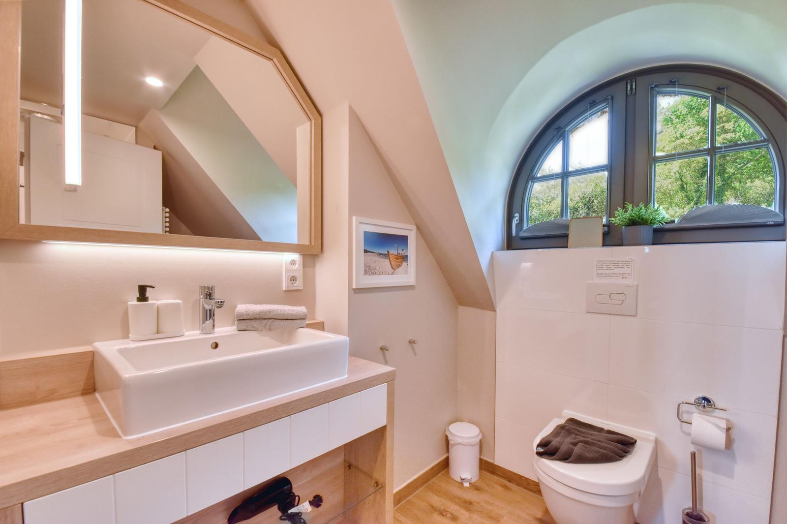 Badezimmer mit Waschbecken, Spiegel und Toilette unter einem Bogenfenster.