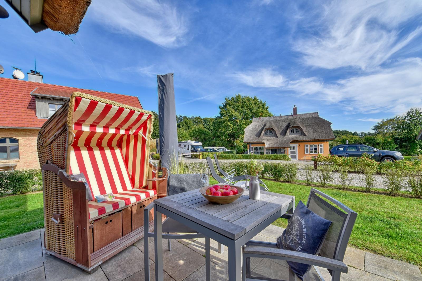 Terrasse mit Strandkorb, Tisch und Stühlen. Im Hintergrund Haus und Grünfläche.