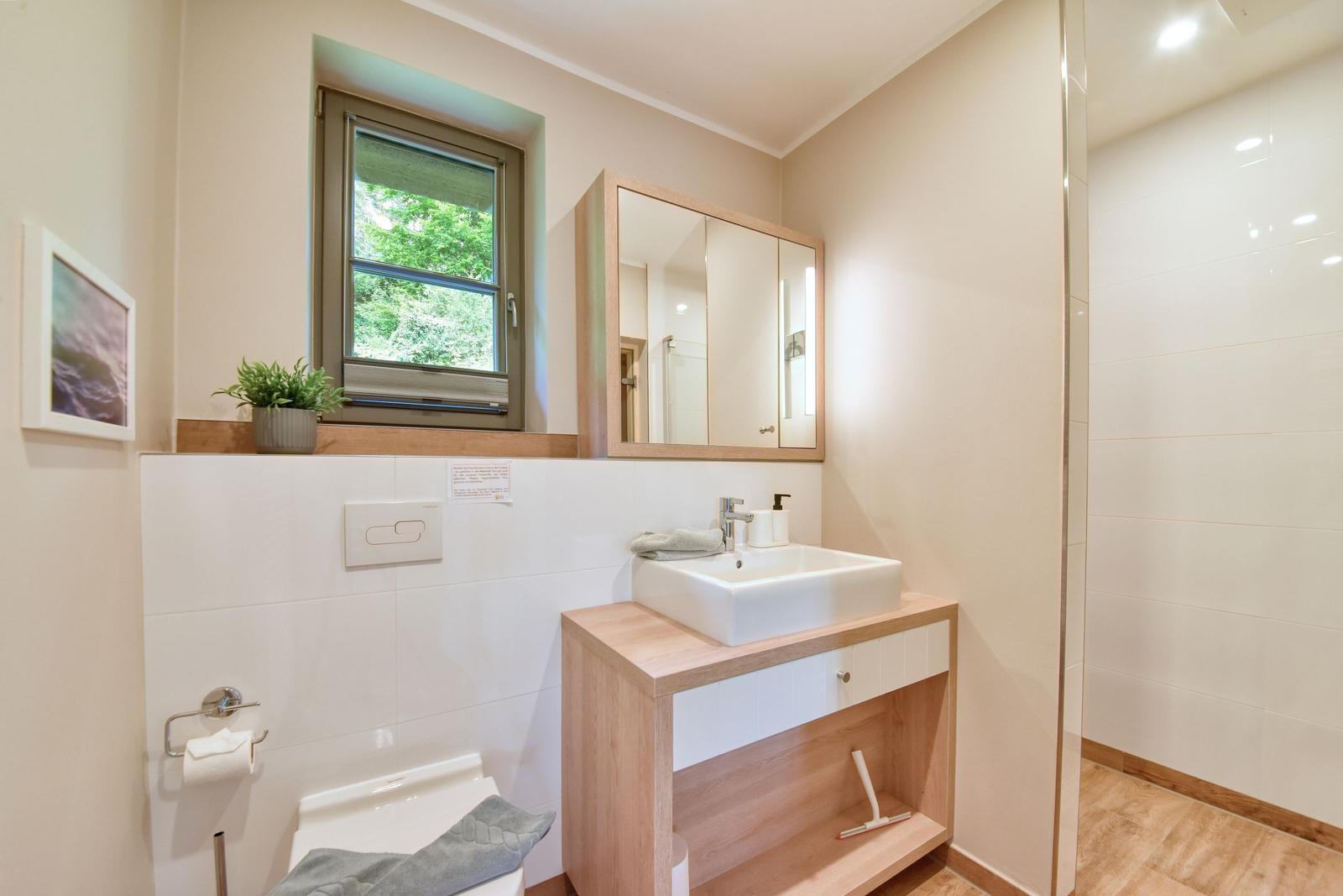 Badezimmer mit Waschbecken, Spiegel und Fenster mit Blick auf Grün.