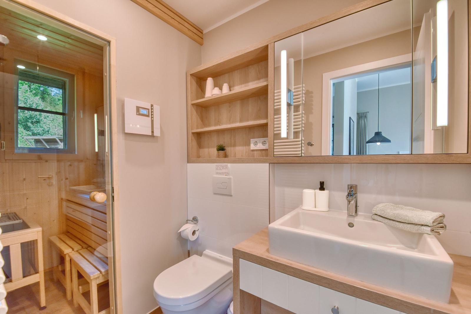 Badezimmer mit Dusche, Waschbecken und Toilette. Holzregale und Spiegel mit Beleuchtung.