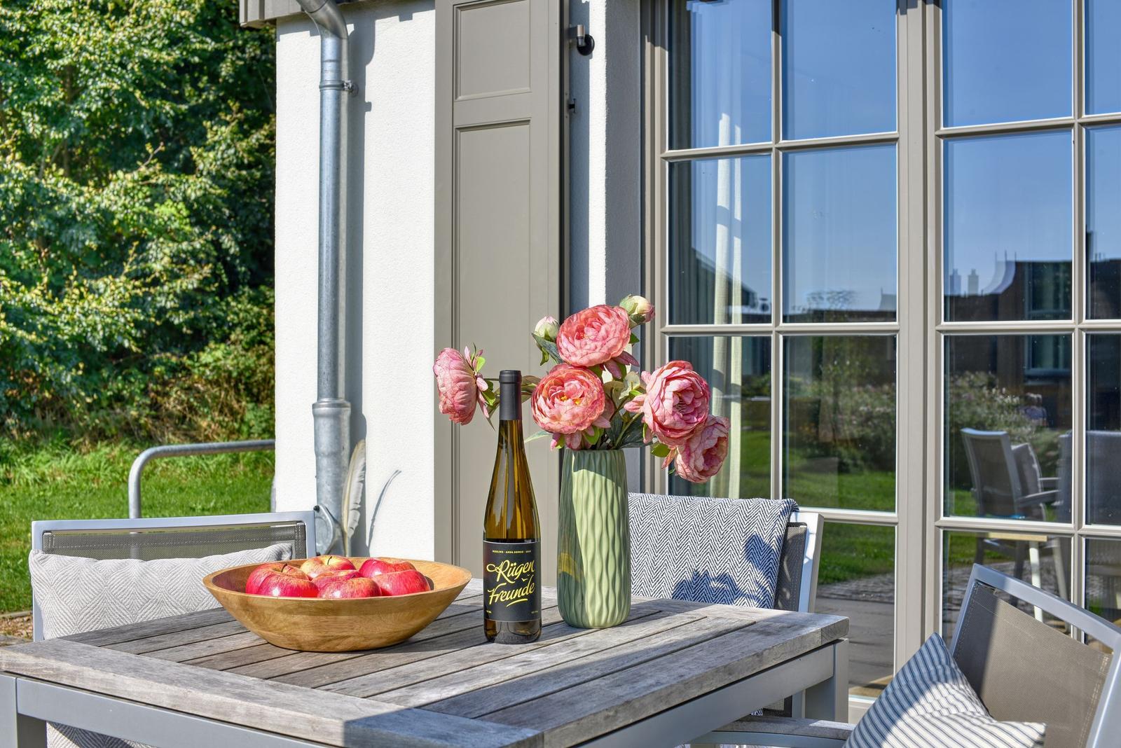 Terrasse mit Tisch, Stühlen, Obstschale, Weinflasche und Blumenarrangement.